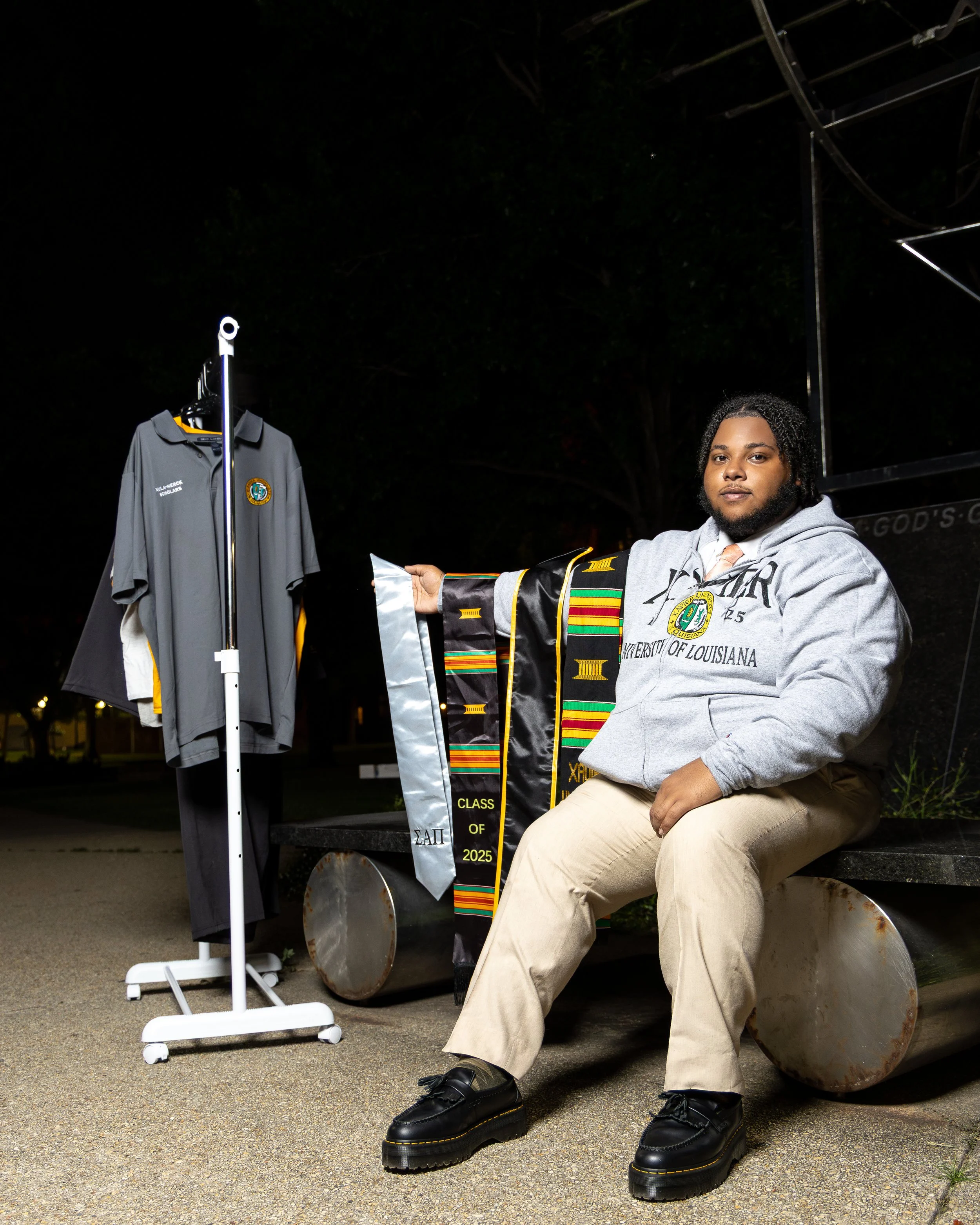 A young man sitting on a park bench at night, wearing a gray hoodie with 'University of Louisiana' written on it, and holding a stole for graduation. Next to him is a small display with additional graduation stoles and a rack with a graduation gown, 