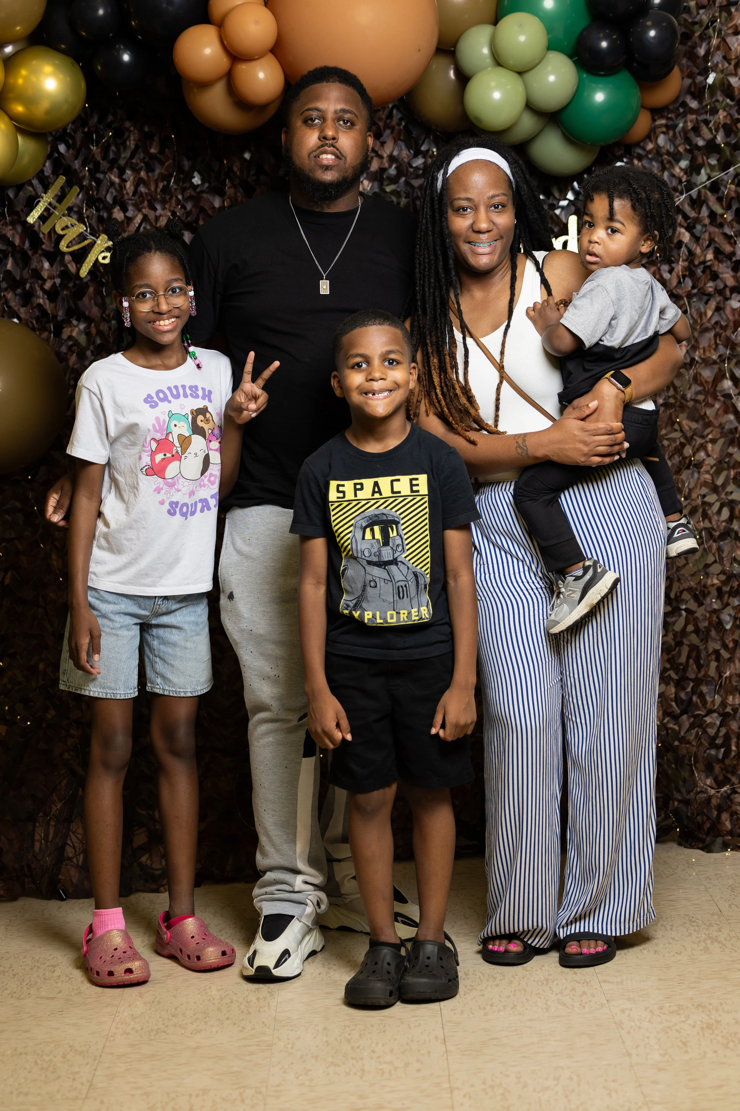 A family of 5 posing in front of a decorative wall with balloons and a banner that reads 'Happy'.