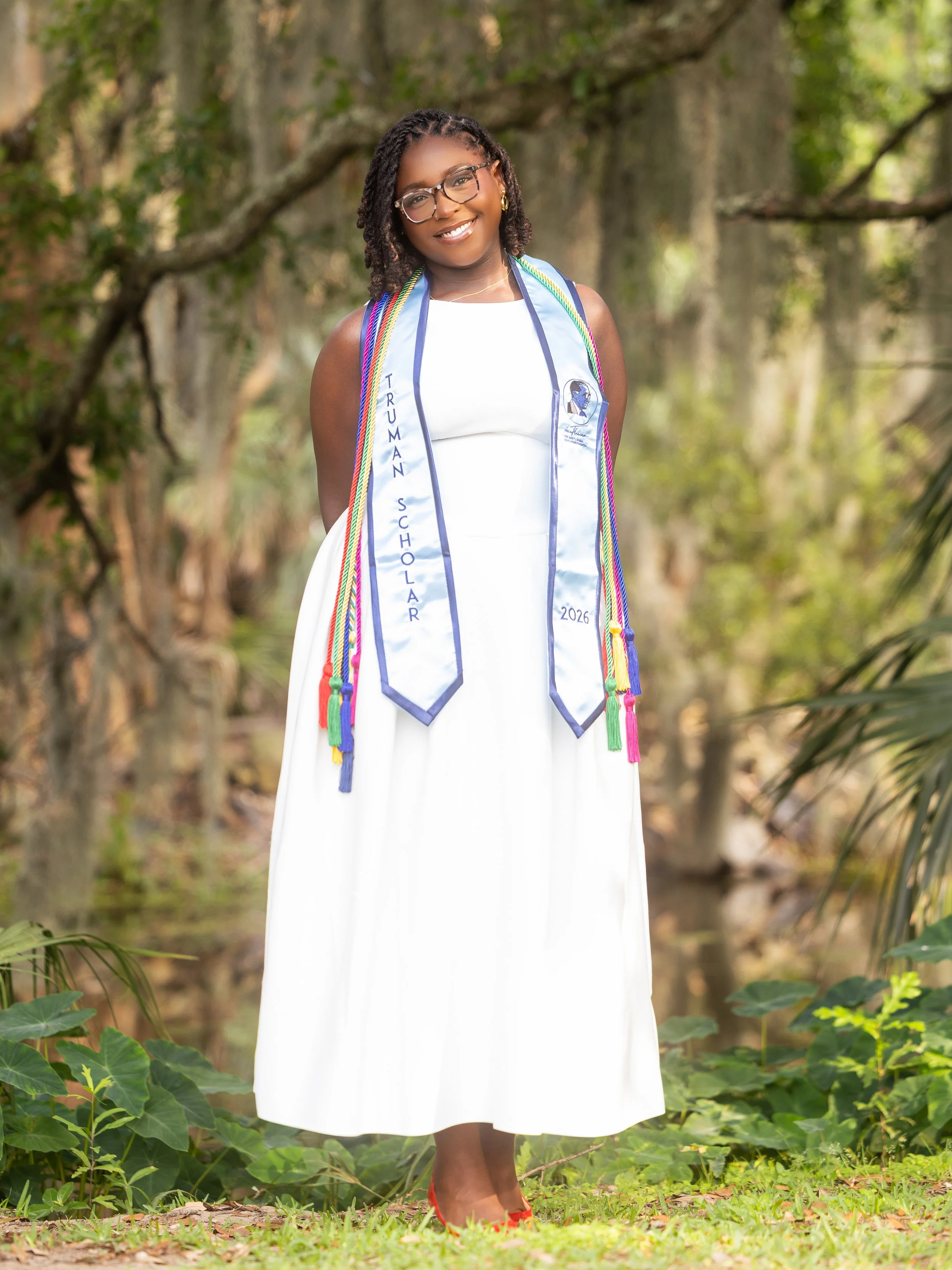 Woman standing outdoors in a forest, wearing a white dress, colorful graduation stole that reads 'TRUMAN SCHOLAR 2026', and red shoes.