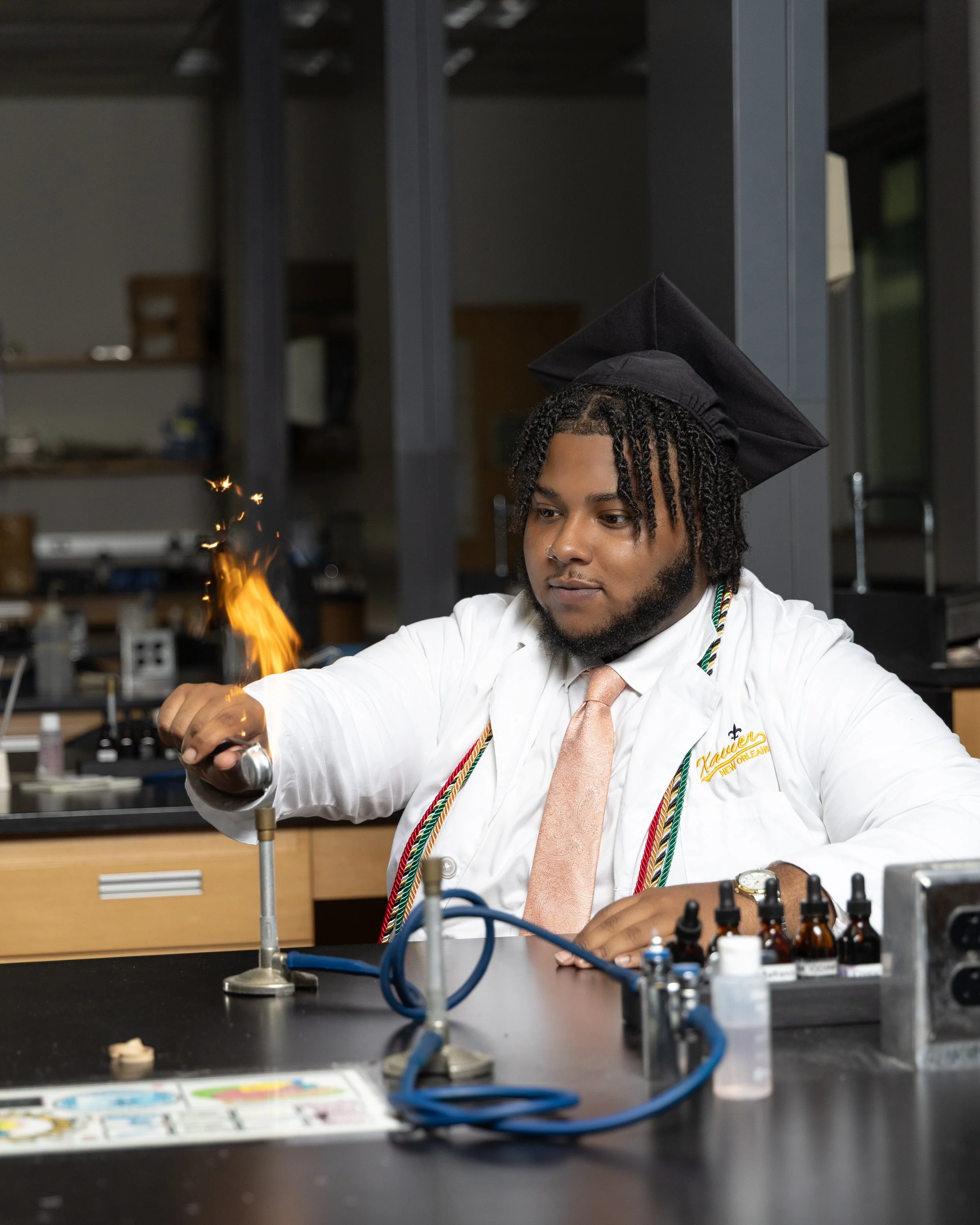 A young man in a white shirt, pink tie, and graduation cap performing a science experiment with a Bunsen burner producing a flame in a laboratory.