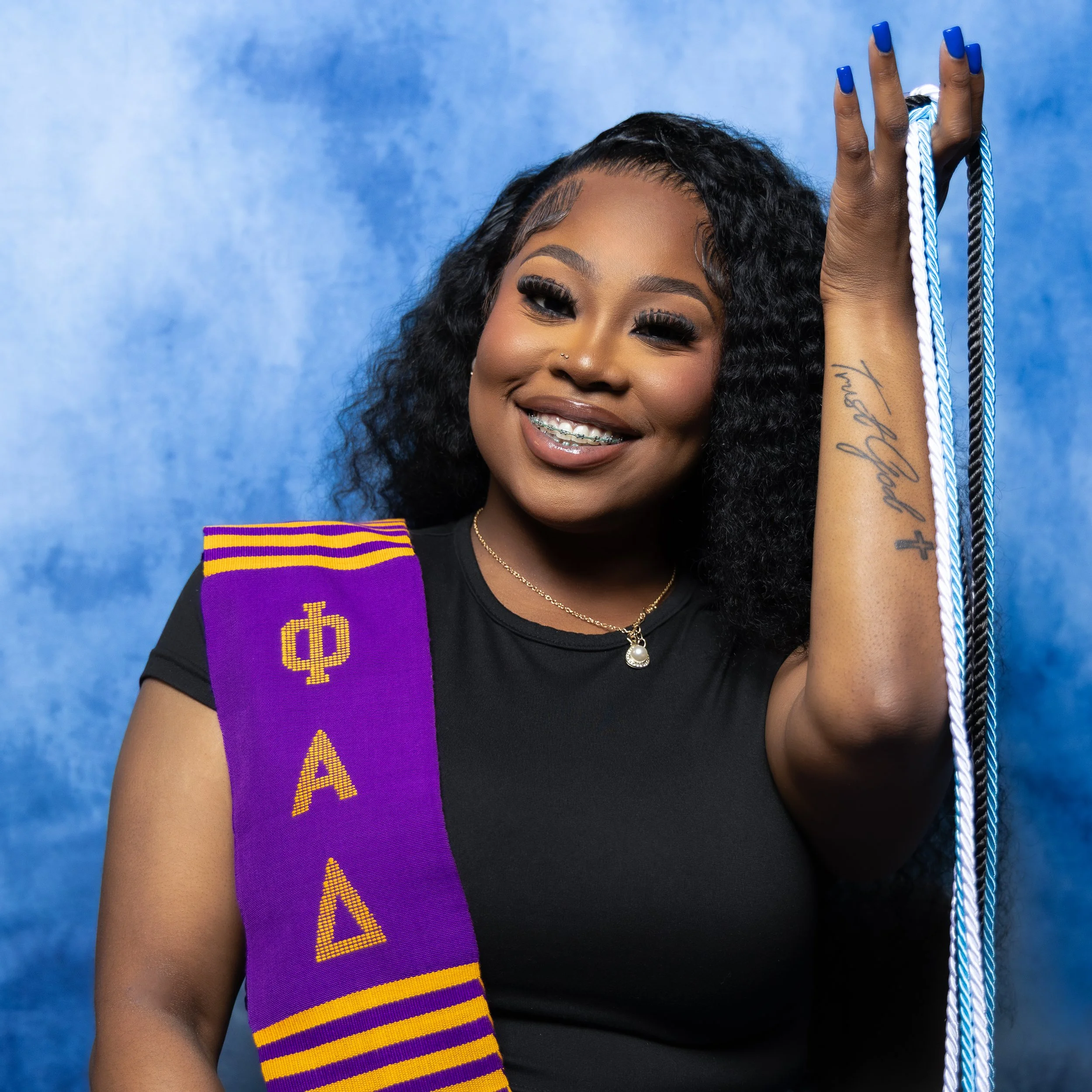 A young woman with curly black hair, smiling and showing braces, wearing a black top. She has blue nails, a tattoo on her arm, and a necklace with a pearl pendant. She is holding graduation cords and wears a purple and gold sash with Greek letters, standing against a blue background.