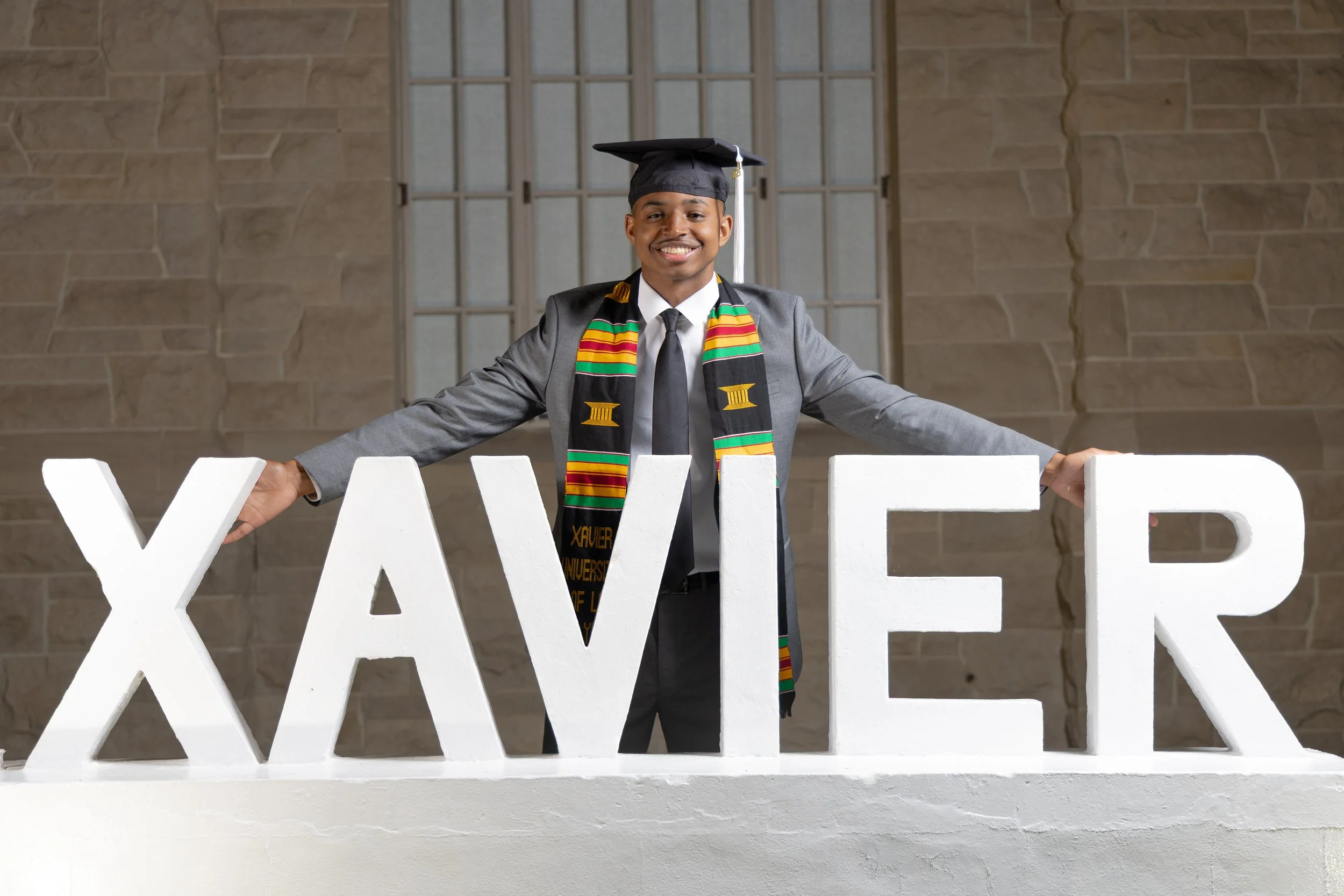 Graduate in cap and gown smiling, standing behind large white letters spelling 'XAVIER' on a white platform, in front of a brick wall.