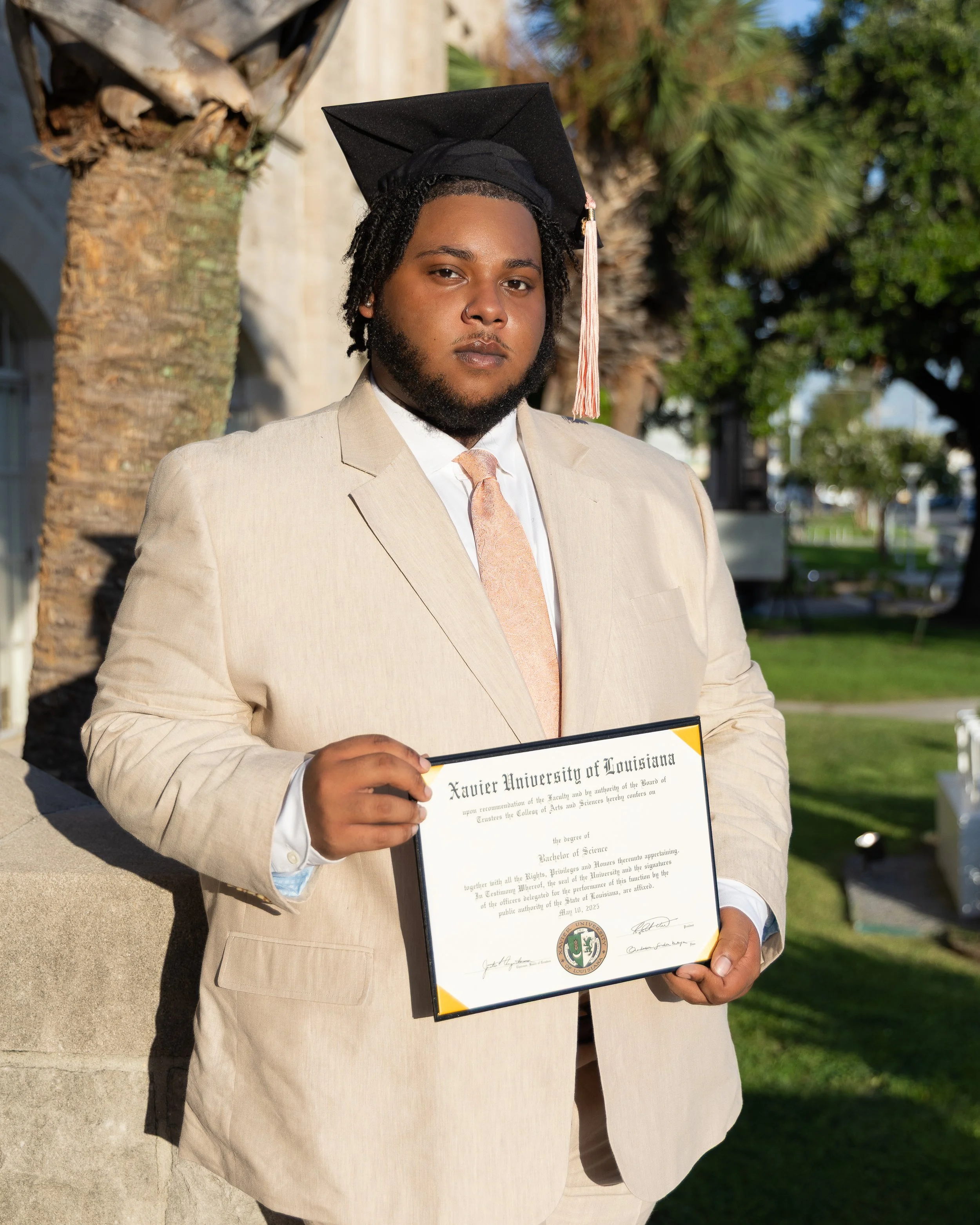Young man in a light beige suit and pink tie, wearing a black graduation cap, holding a diploma from Xavier University of Louisiana, outdoors with palm trees and greenery.