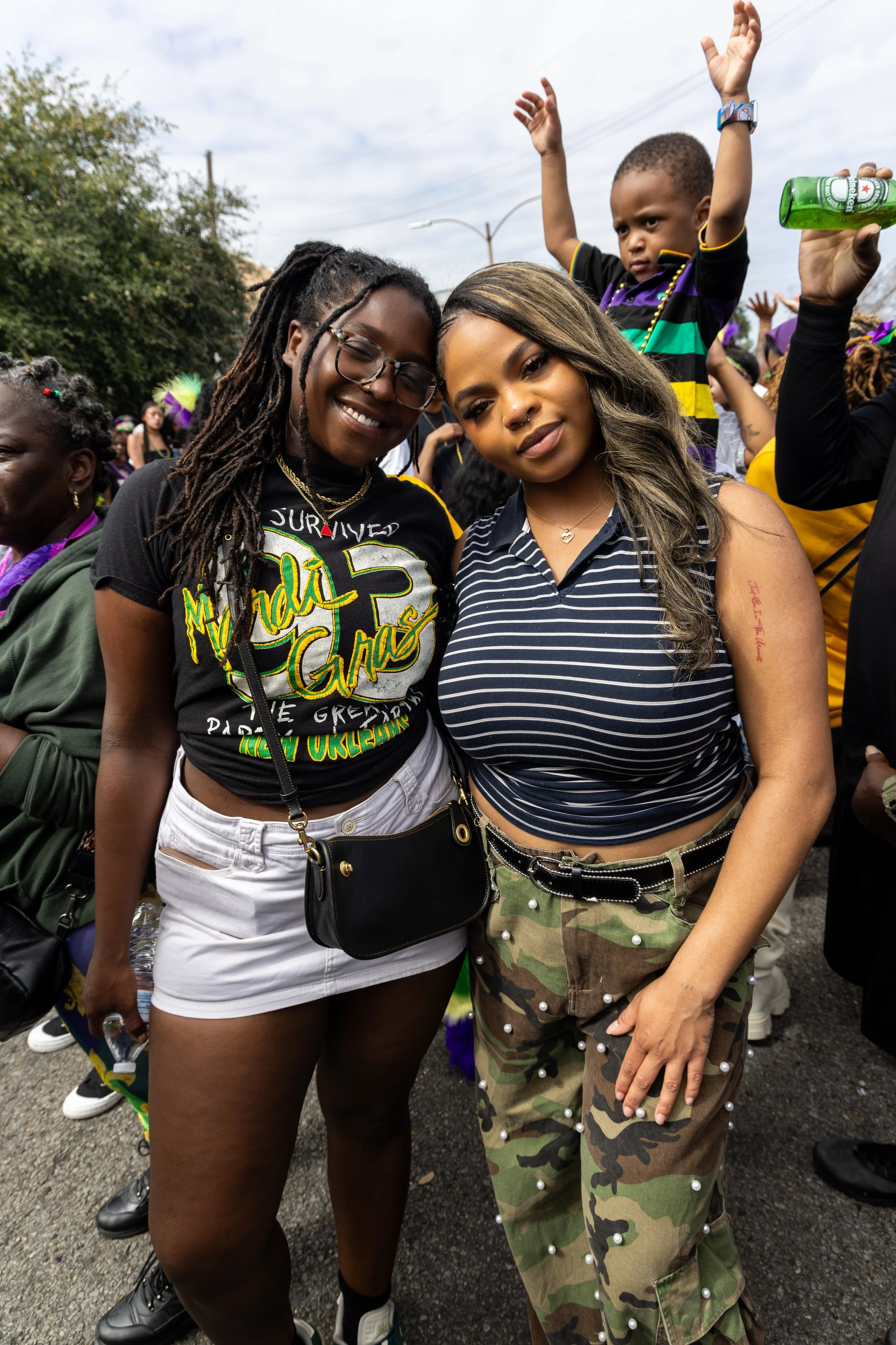 Two women standing close together at a parade or outdoor event, one with dreadlocks and glasses wearing a black shirt with colorful text, and the other with wavy hair wearing a navy and white striped shirt and camouflage pants with pearl embellishmen