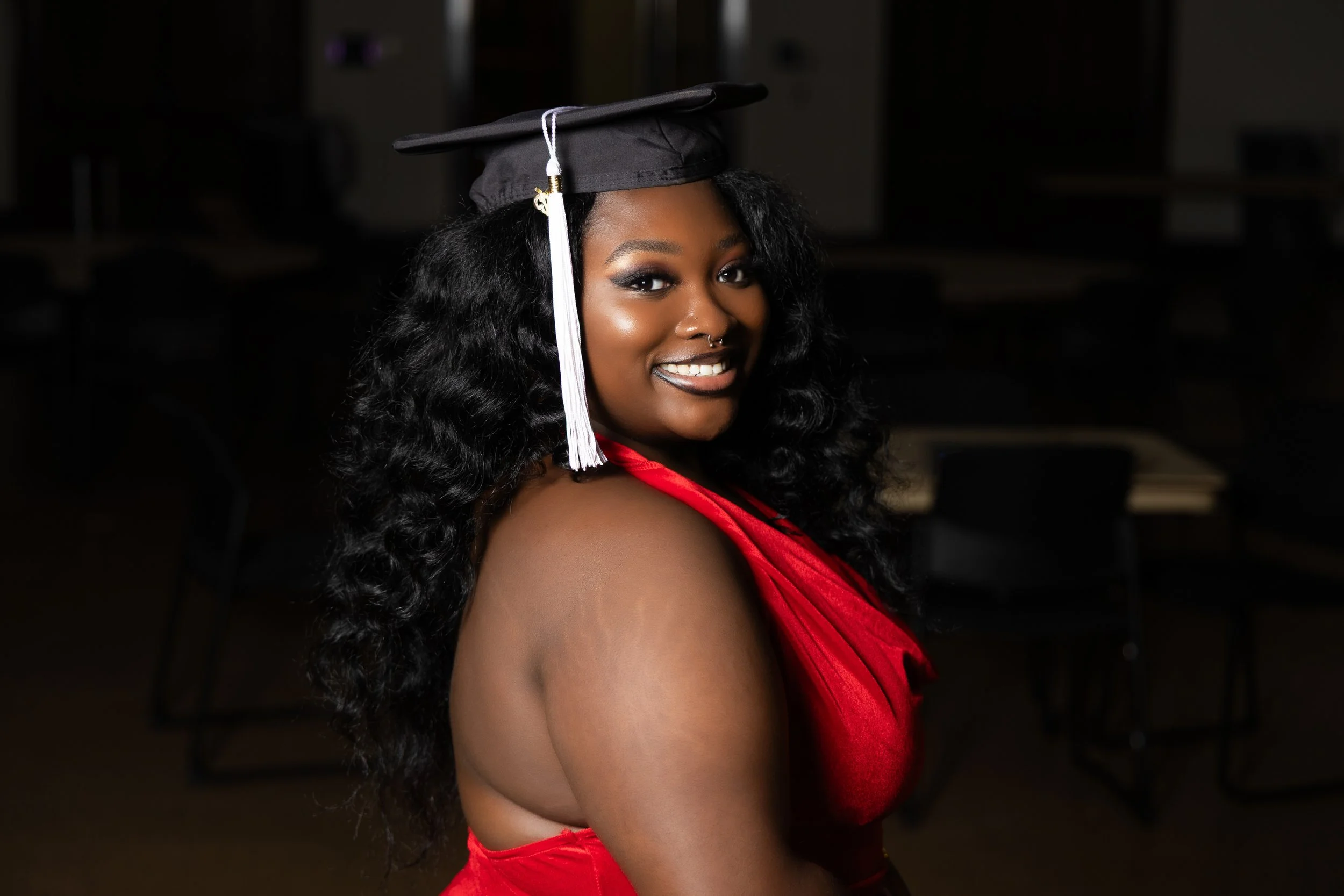 A young woman wearing a graduation cap with a white tassel and a red satin gown, smiling for a portrait inside a dimly lit room.