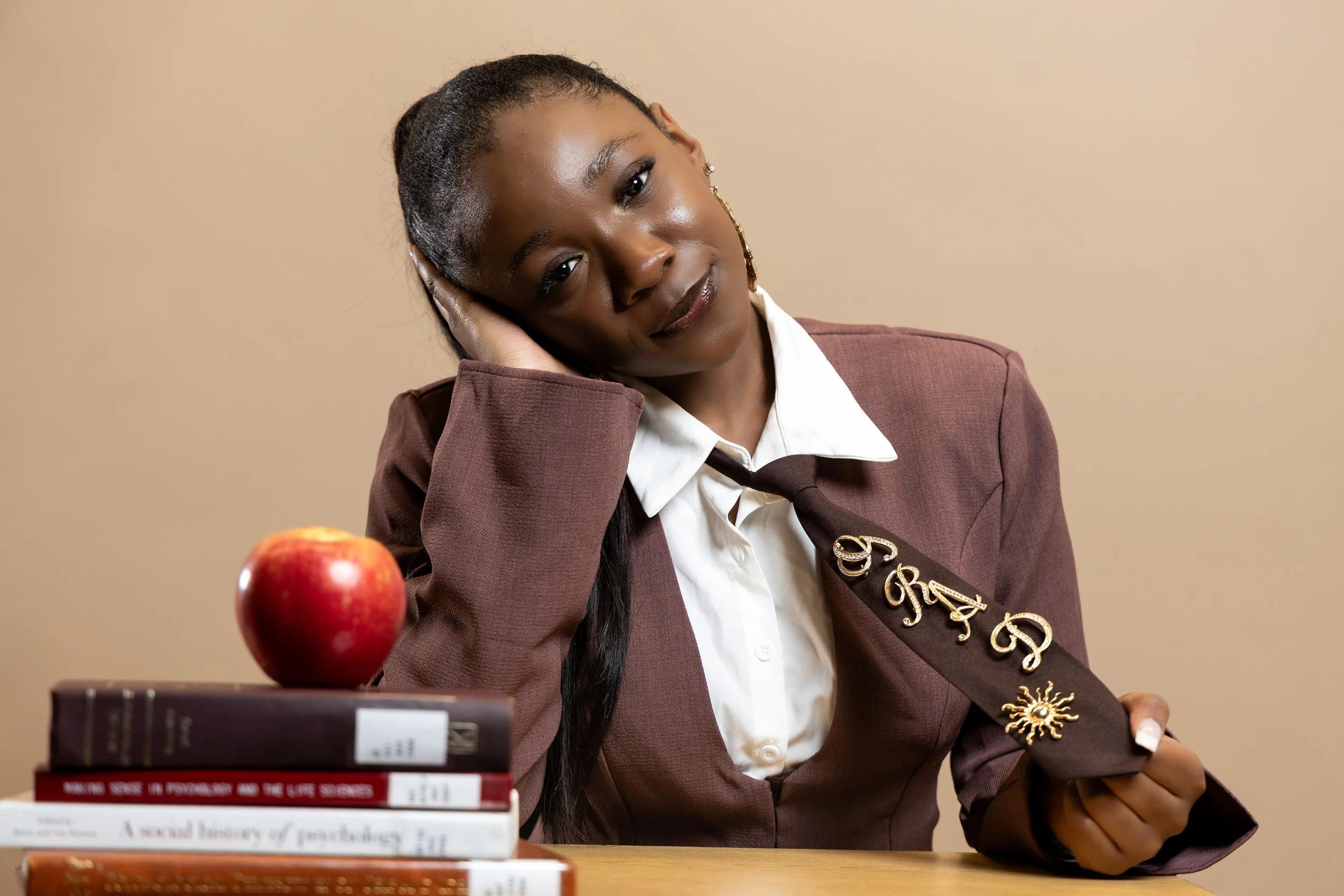 A woman in a brown blazer and white shirt resting her head on her hand at a desk, holding a brown tie with embroidered gold letters and a sun design, with a stack of books and a red apple on the desk.