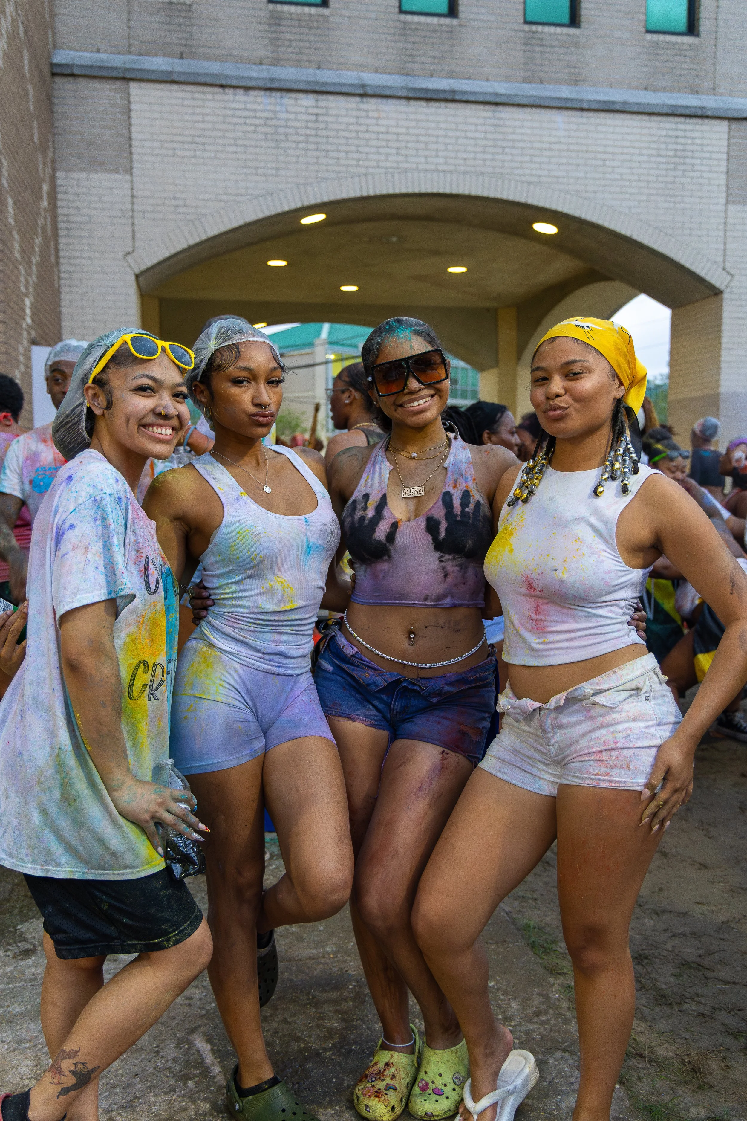 Four women posing together at a color run event, covered in colored paint and wearing casual, colorful outfits.