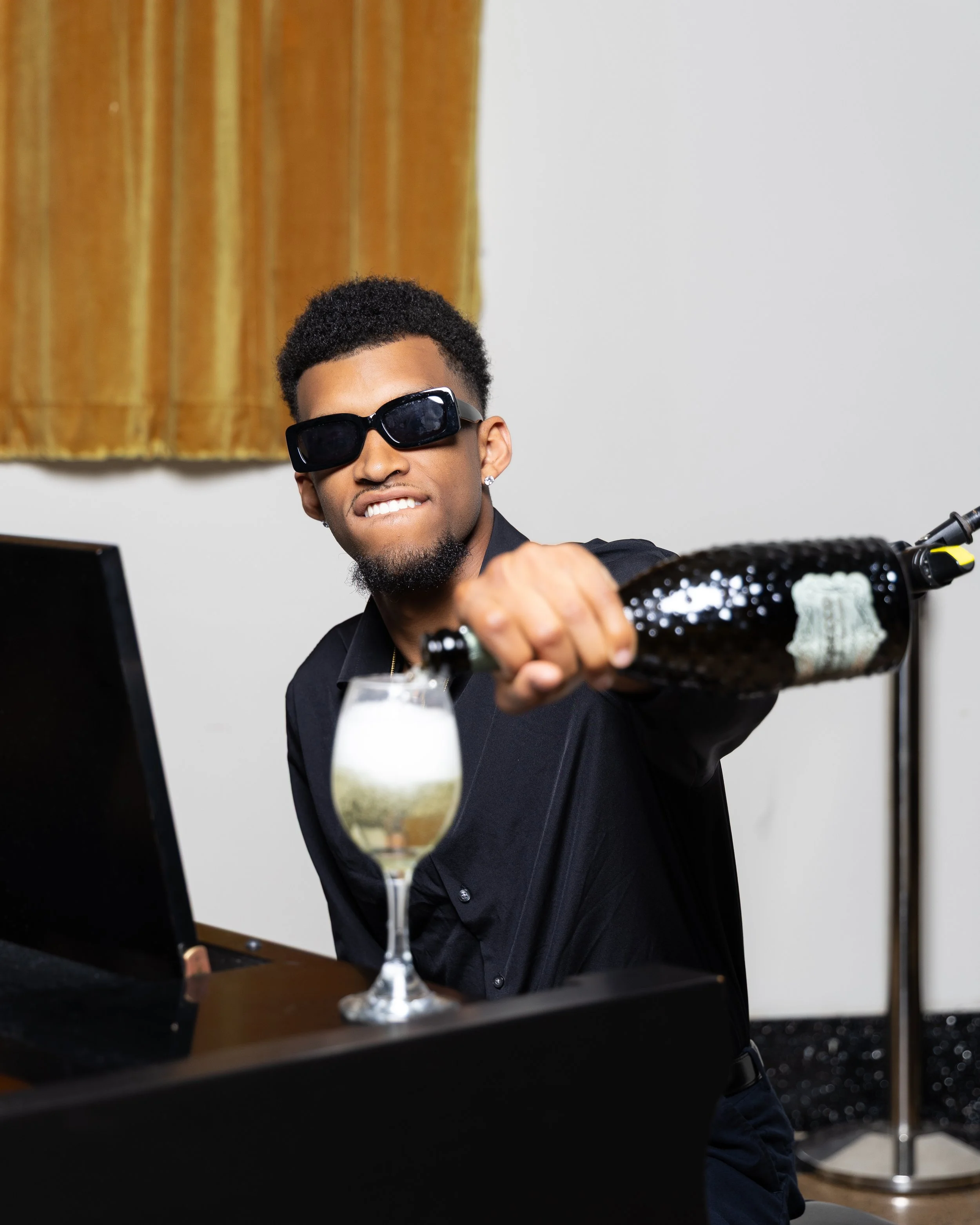 Young man wearing sunglasses is pouring champagne into a glass on top of a piano.