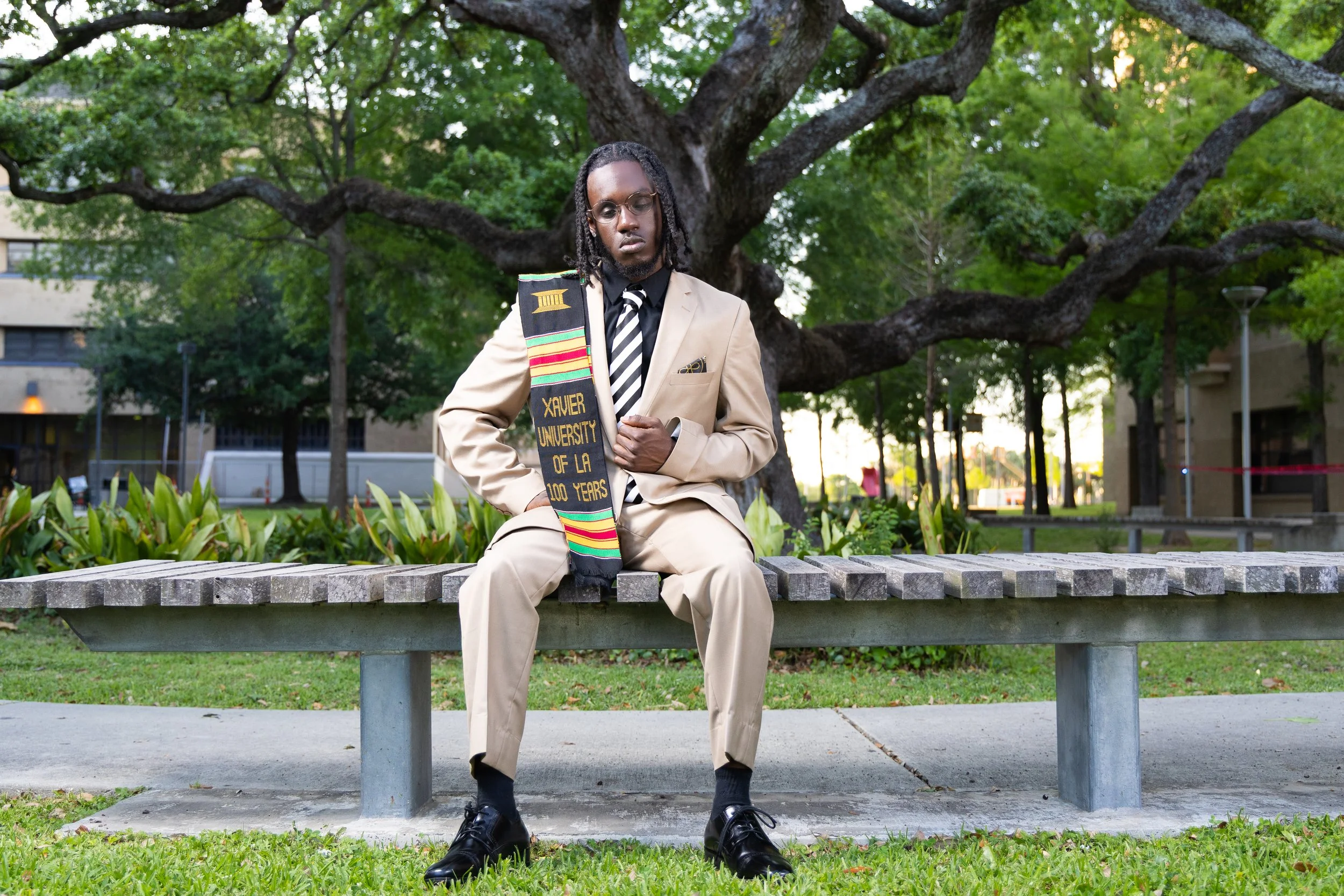 A young man in a beige suit and black shirt sits on a park bench under a large tree, holding a graduation stole that reads 'Xavier University of LA 100 Years'.