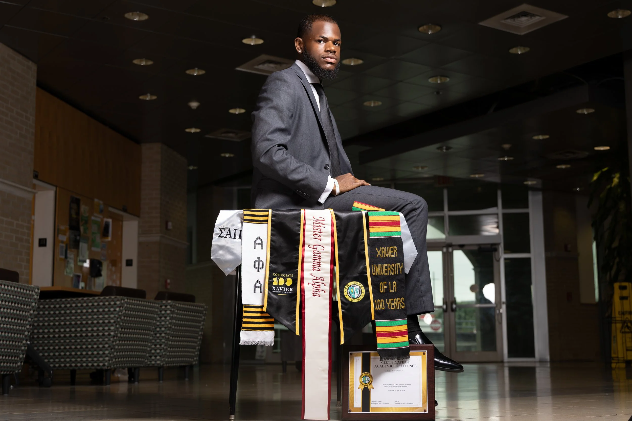 A man in a gray suit sitting on a table covered with graduation honor cords, stoles, and a framed certificate in an indoor setting.