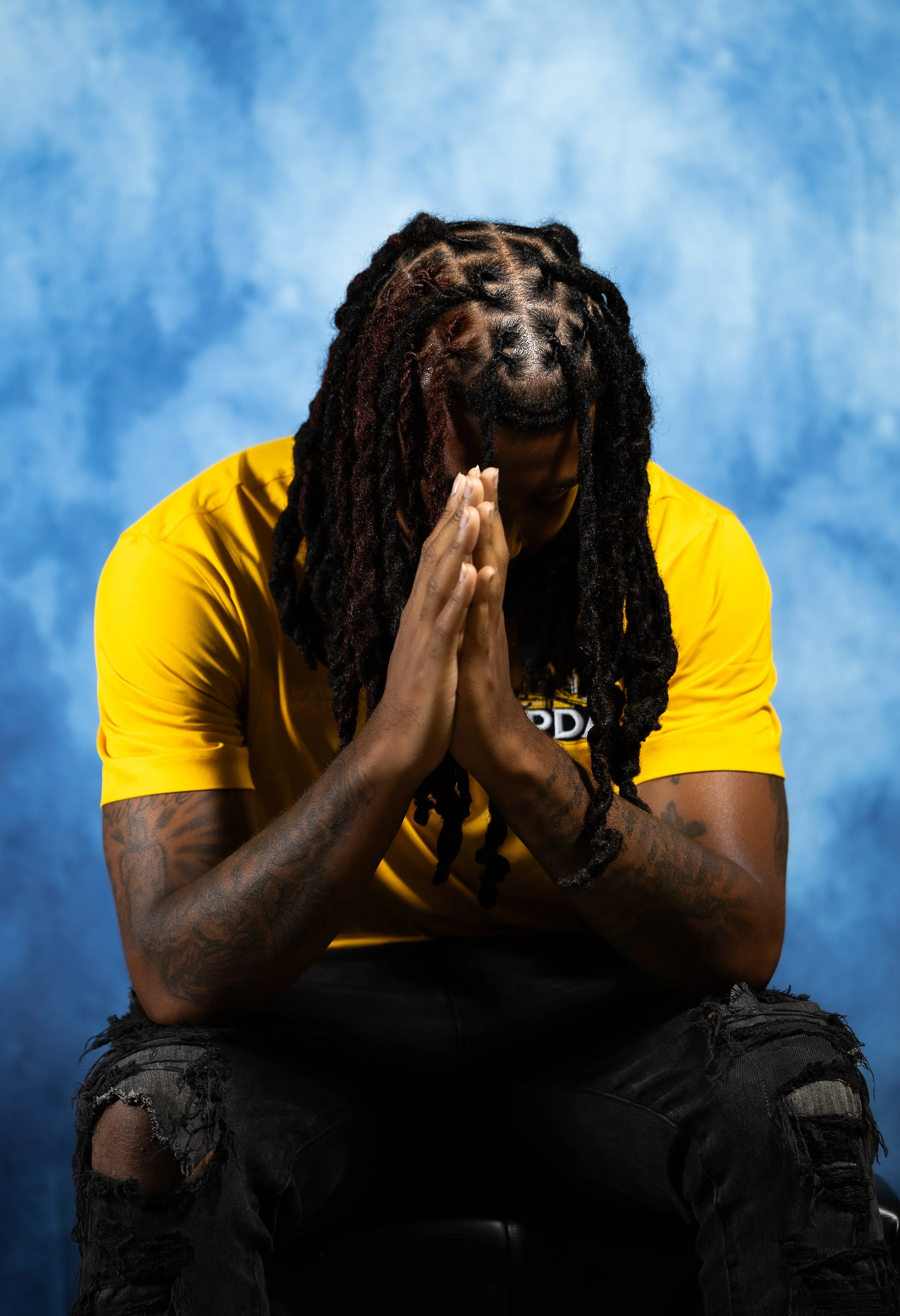 A person with long locs wearing a yellow shirt, sitting with hands pressed together in a prayer or reflection pose, against a blue background.