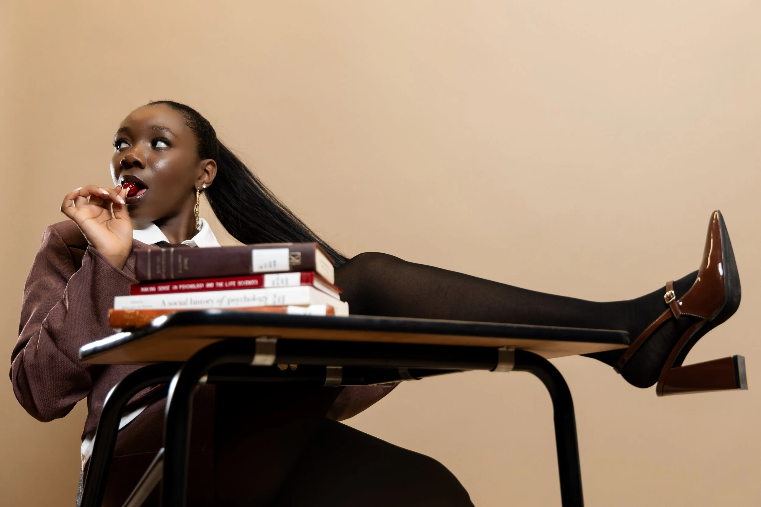 A woman with dark skin and long black hair, dressed in a brown blazer, white shirt, and black tights, lies on a desk with her legs elevated on the surface. She is holding a red lollipop in her mouth and has a stack of books on the desk in front of he