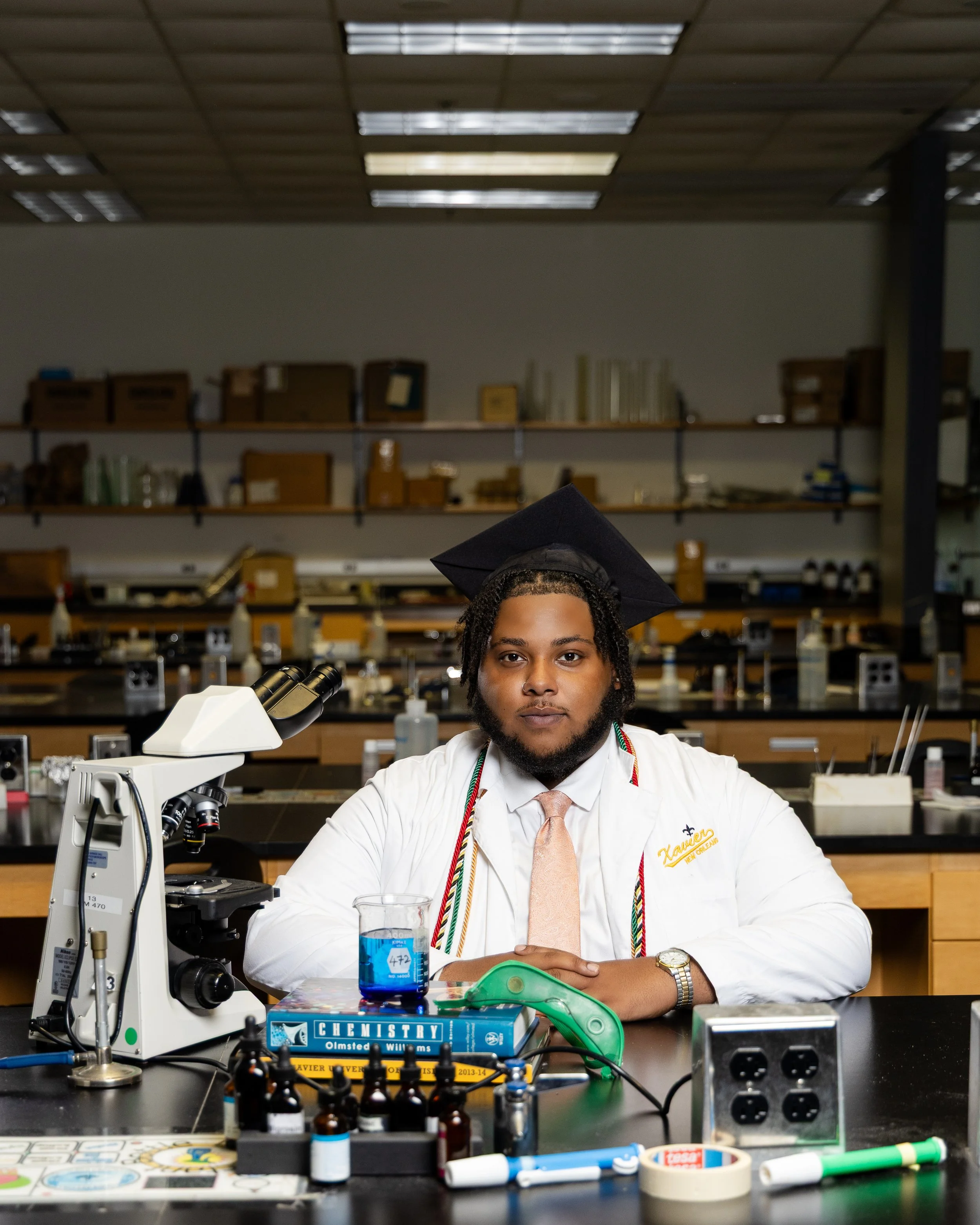 A young man wearing a graduation cap and white lab coat in a science laboratory, seated at a lab table with scientific equipment including a microscope, chemistry books, glass beakers, and scientific supplies. 