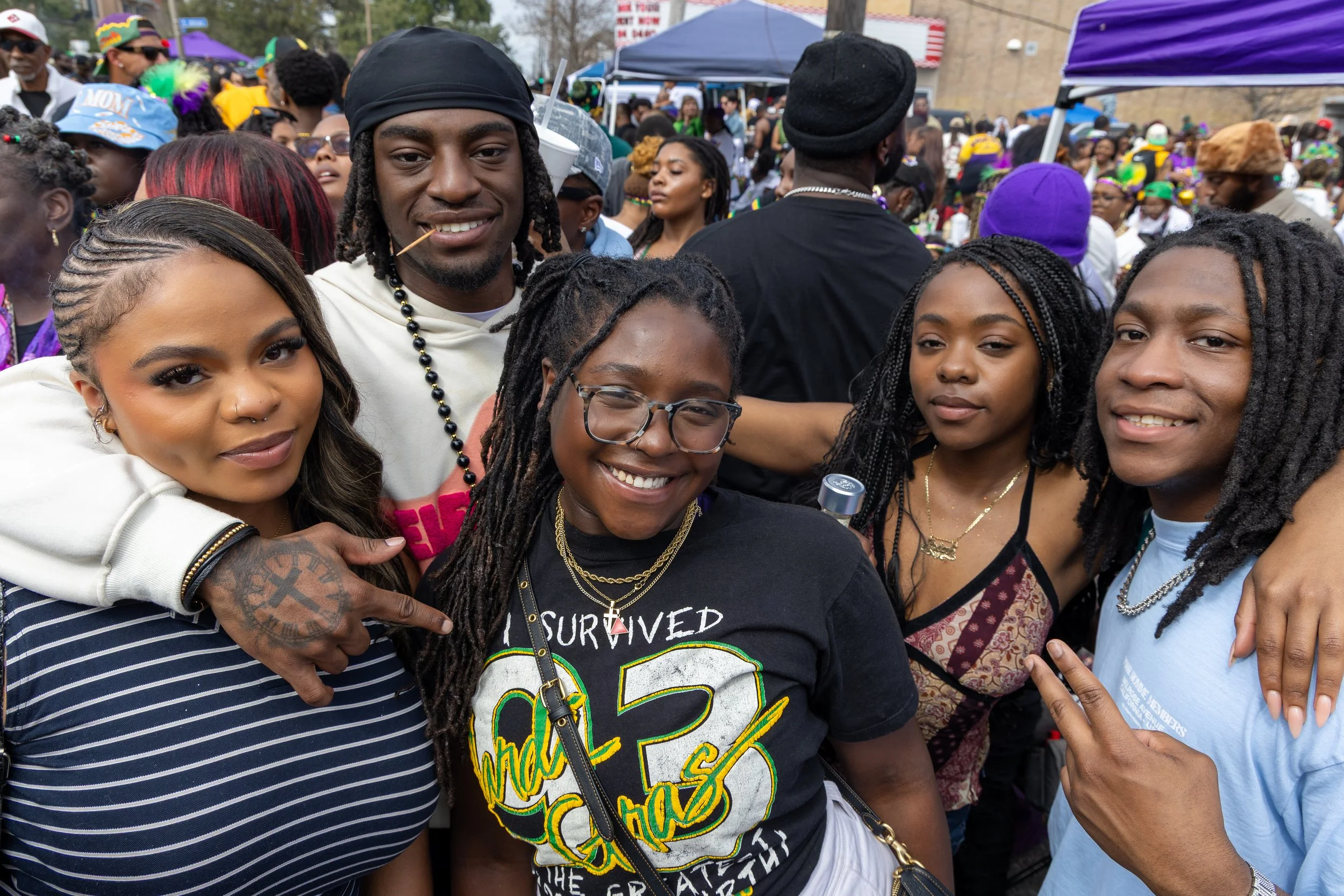A diverse group of people gathered outdoors at a festival or parade, some wearing colorful clothing and accessories, with tents and a crowd in the background.