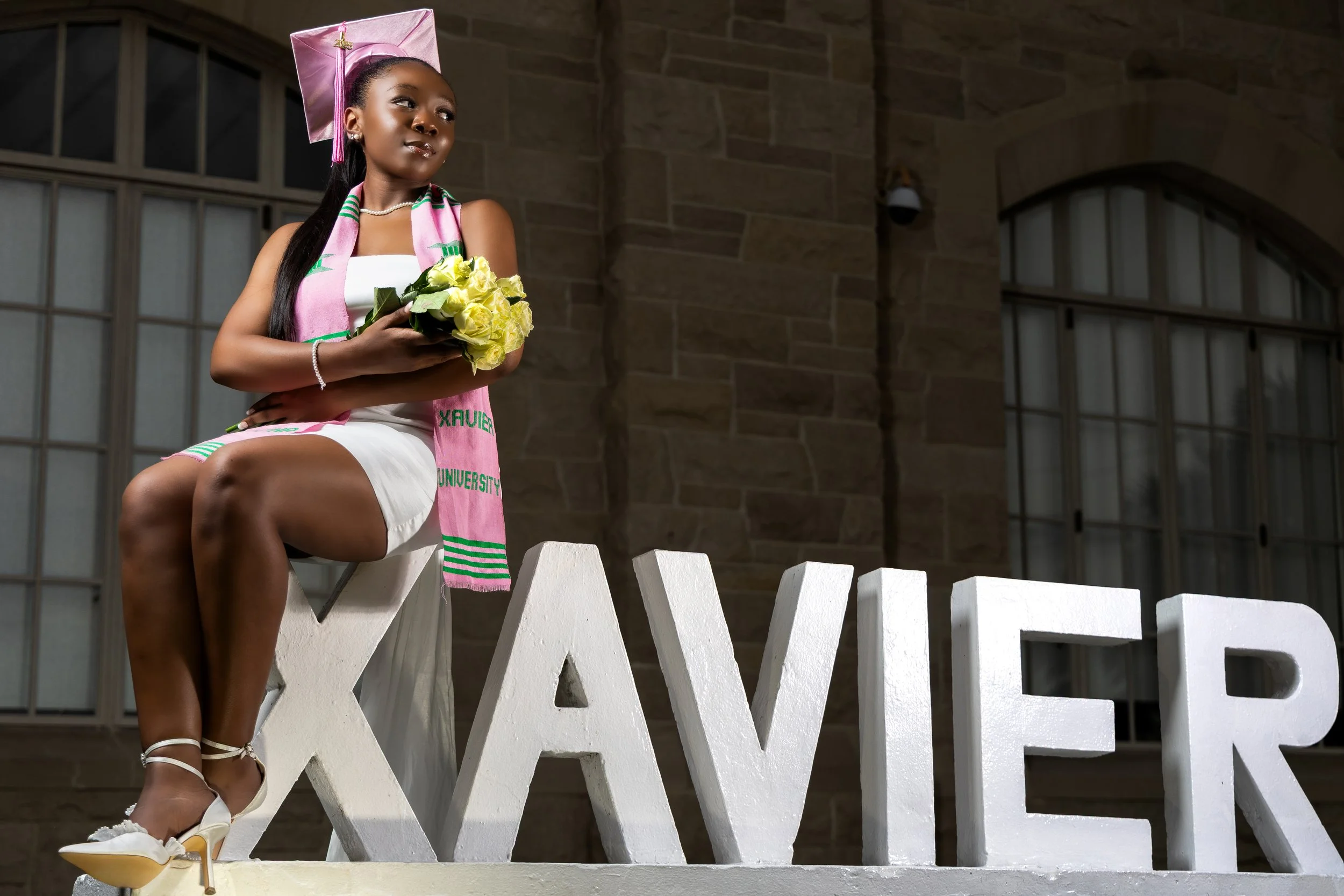 A young woman in graduation attire, sitting on the letter 'X' in a display that spells 'XAVIER', holding a bouquet of yellow flowers, wearing a pink graduation cap and sash, in front of a stone building with large windows.
