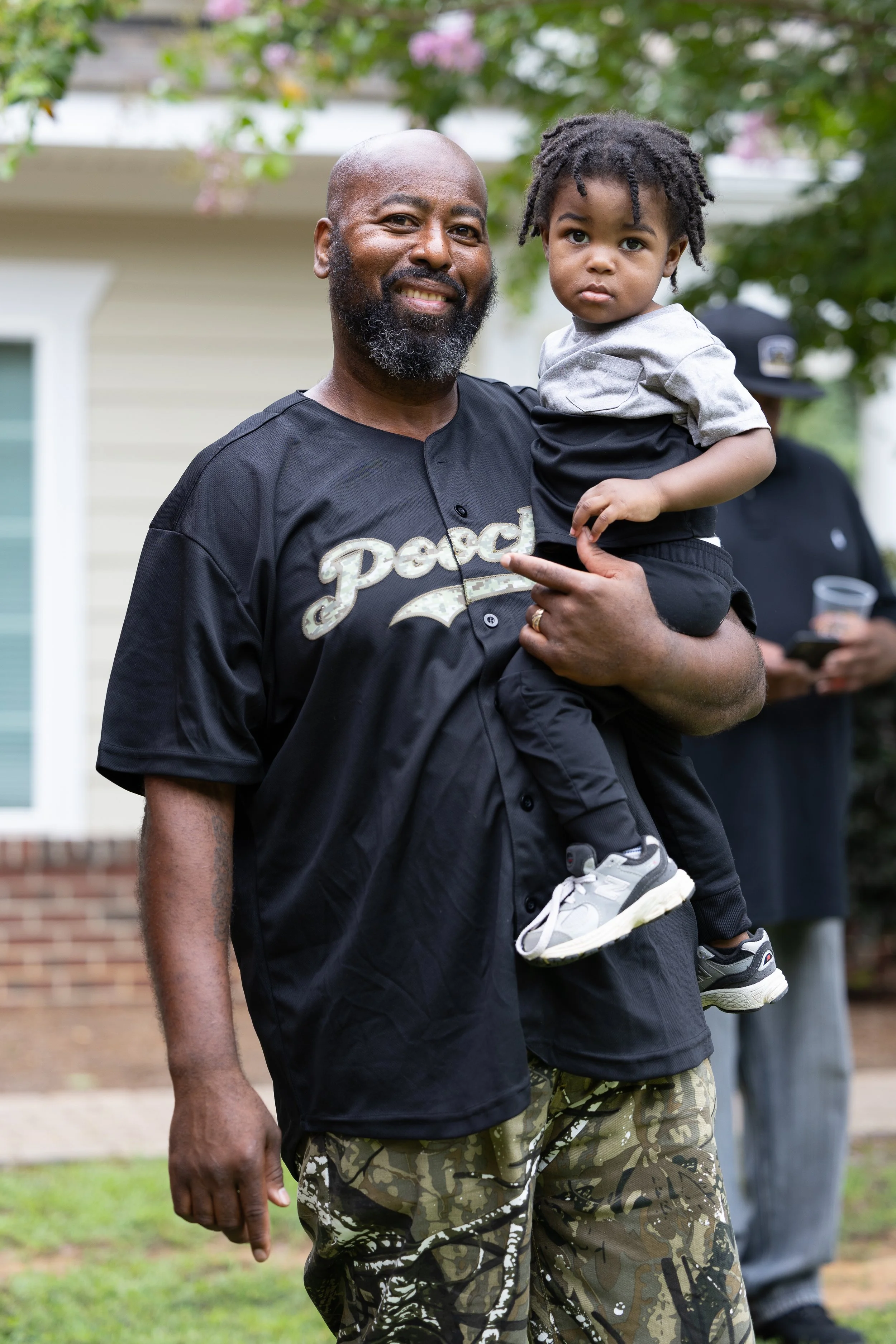 A smiling man with a beard holding a young child in an outdoor setting, with greenery and a house in the background.