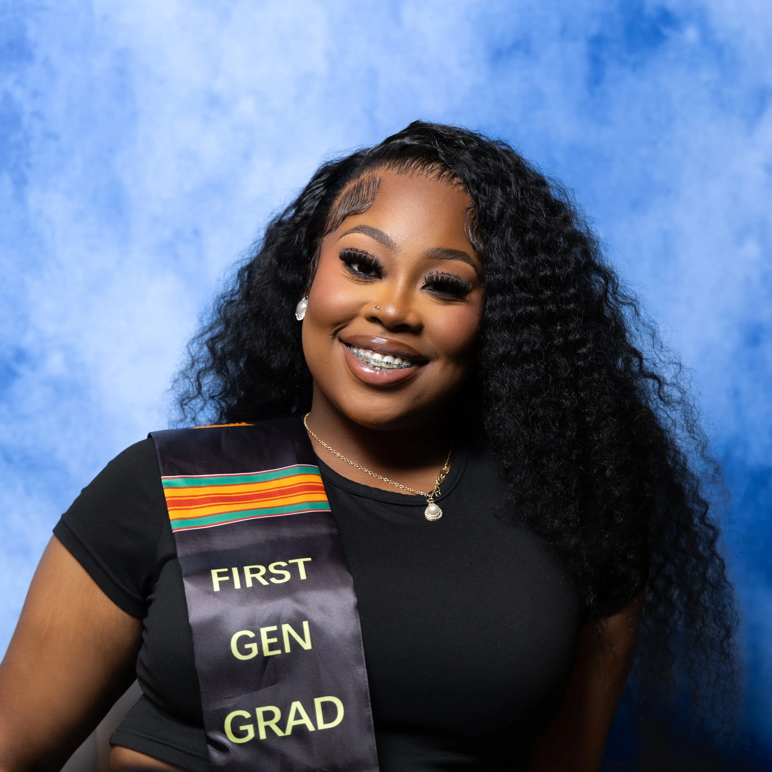 A young woman with curly black hair, smiling, wearing a black top with a sash that reads 'First Gen Grad,' and a pearl necklace, against a blue background.
