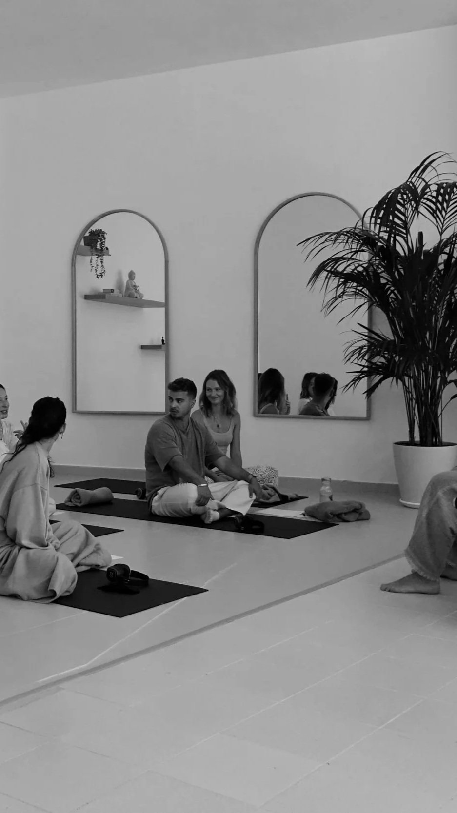 Group of people participating in a yoga or meditation class in a serene, minimalistic room with large mirrors and a potted plant.