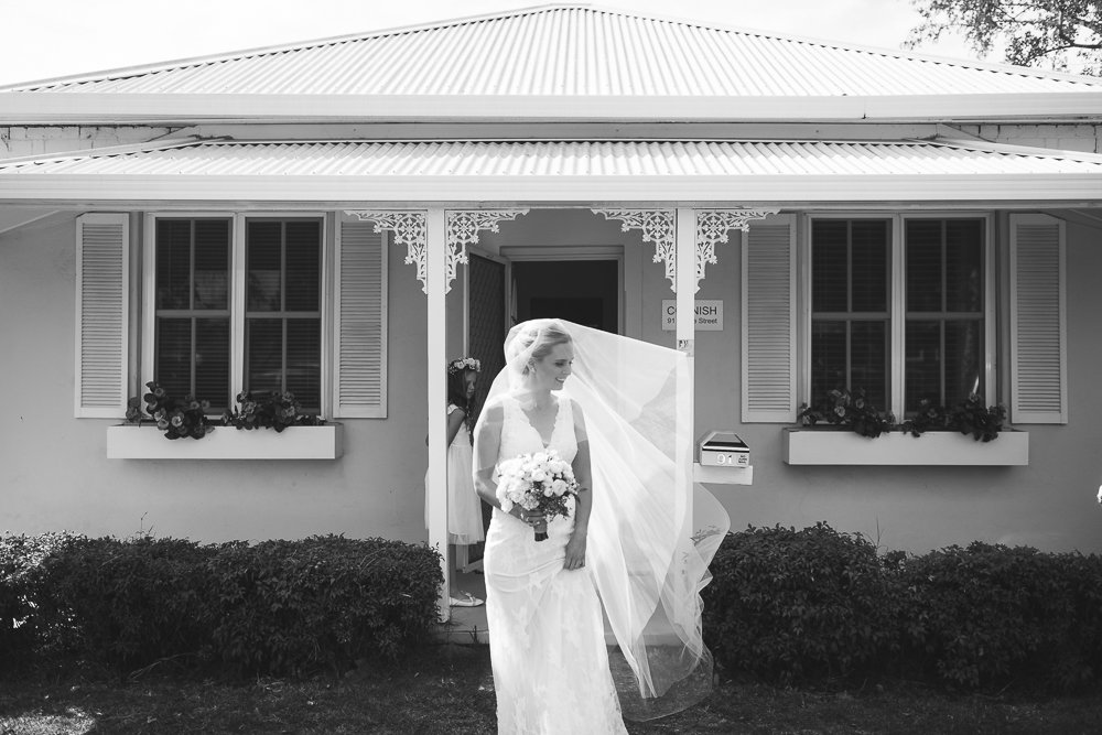 Black and white photograph of a bride in a wedding dress holding a bouquet, standing outside a house with shutters and flower boxes, with a woman in a dress partly visible behind her.