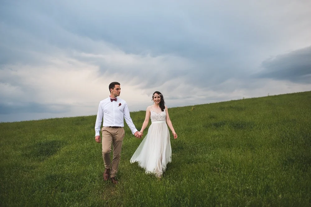 A bride and groom holding hands and walking on a grassy hill against a cloudy sky.