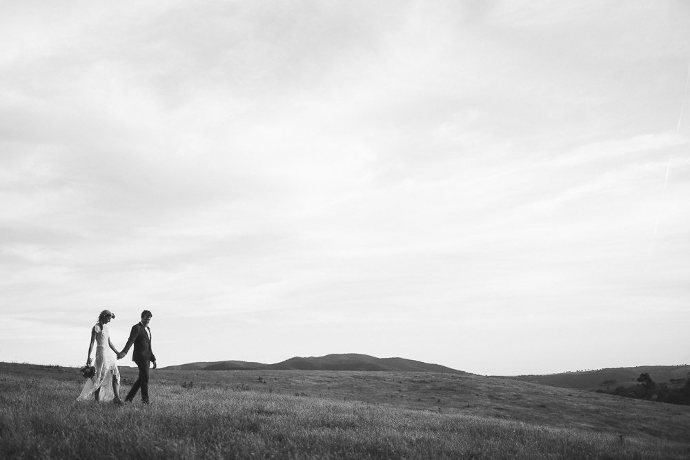Black and white photo of a bride and groom walking hand in hand across a grassy field with distant rolling hills and a cloudy sky.
