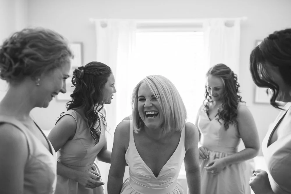 Group of women laughing and smiling in a bright room, dressed in light-colored dresses.