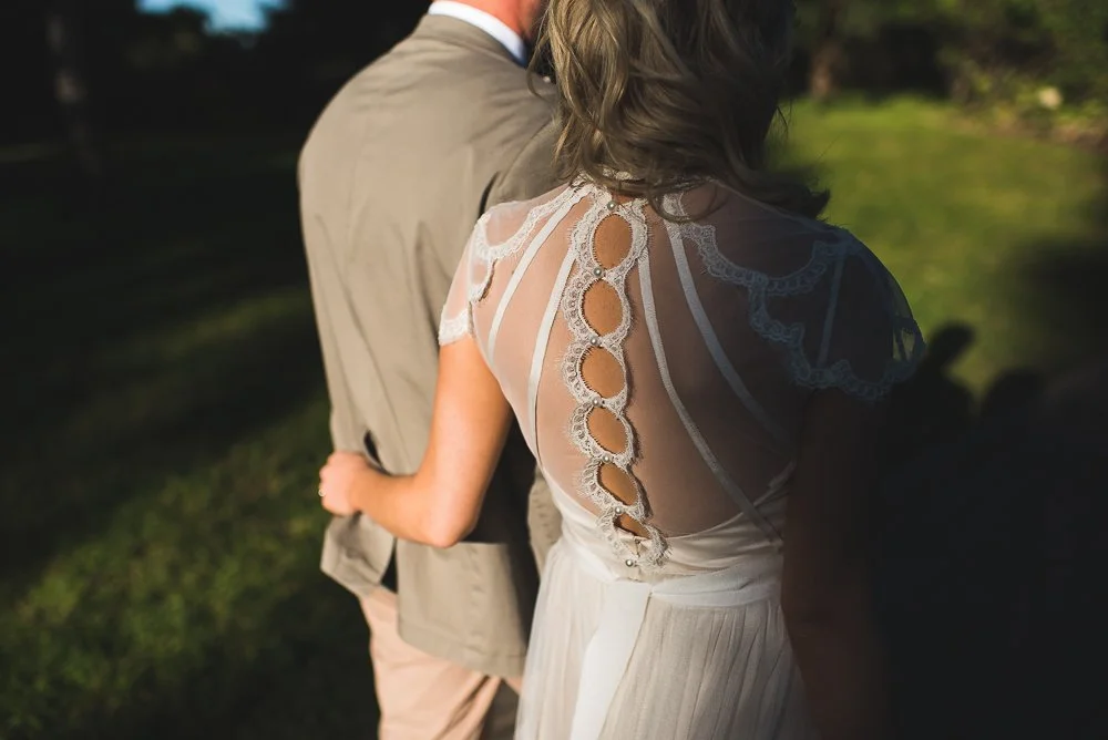 A woman in a white dress with intricate lace detailing on the sleeves, standing close to a man dressed in a beige suit outdoors. The sunlight creates shadows, highlighting the textures of their clothing against a background of green grass and trees.