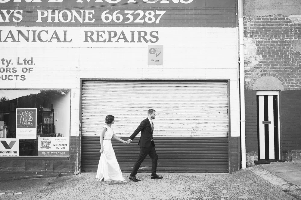 A bride and groom walking hand in hand on a city sidewalk in front of a closed shop with a metal security shutter, a window with signs, and brick walls.