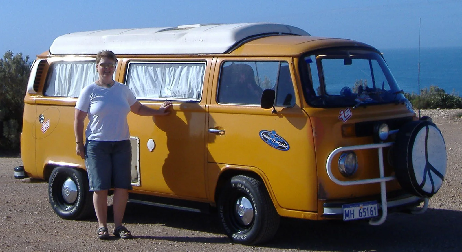 Person standing next to a yellow vintage camper van on a coastal landscape