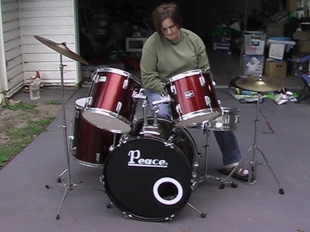 A woman sitting behind a red and black Peace brand drum set on a driveway outside a garage.