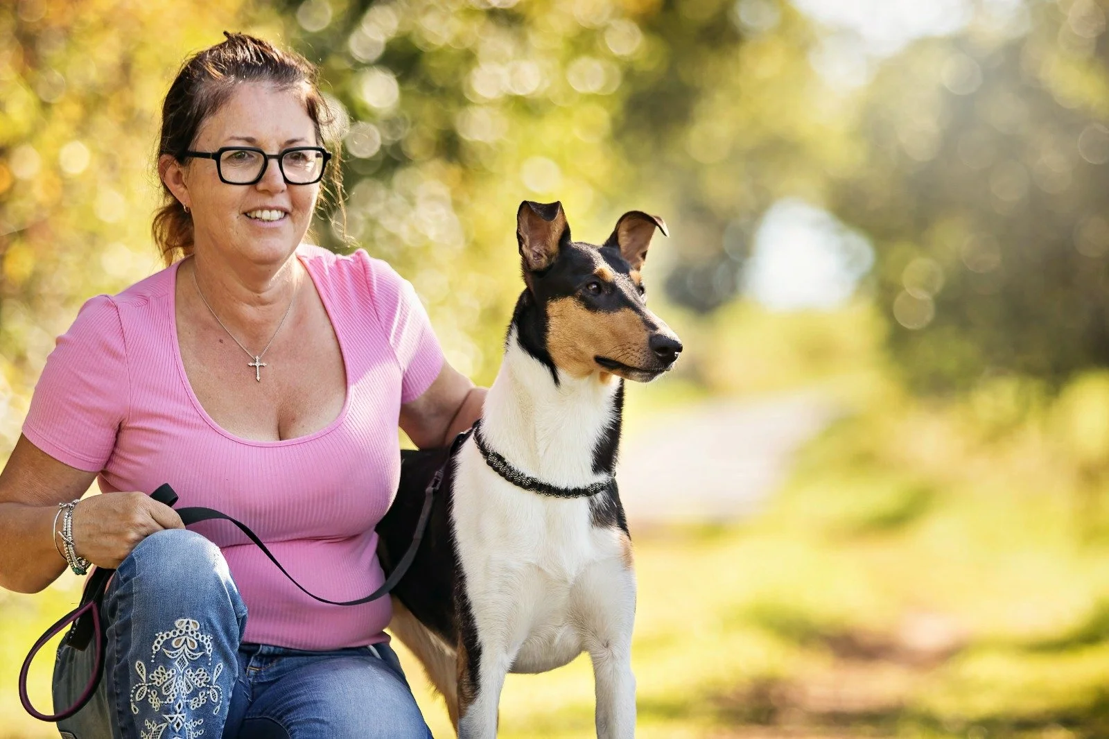 Una donna con occhiali, indossa una maglietta rosa e jeans, è con un Collie Smooth durante una passeggiata all'aperto in un parco autunnale