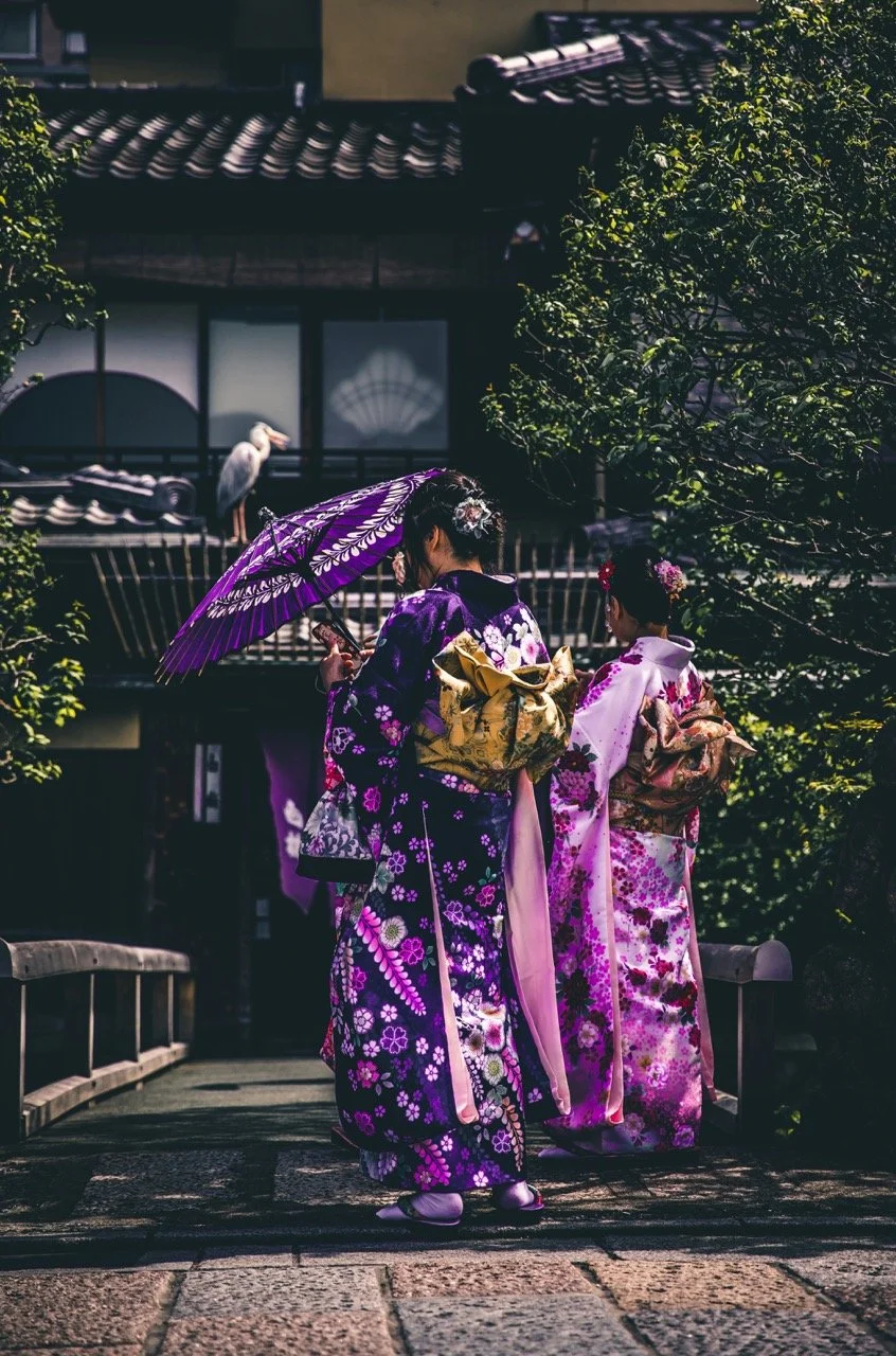 Two women dressed in traditional kimonos walk together on a stone path, with one of them holding a purple parasol. They are near a traditional Japanese building with cedar tiles and a bamboo fence, surrounded by trees. A white crane is visible in the background on a balcony.