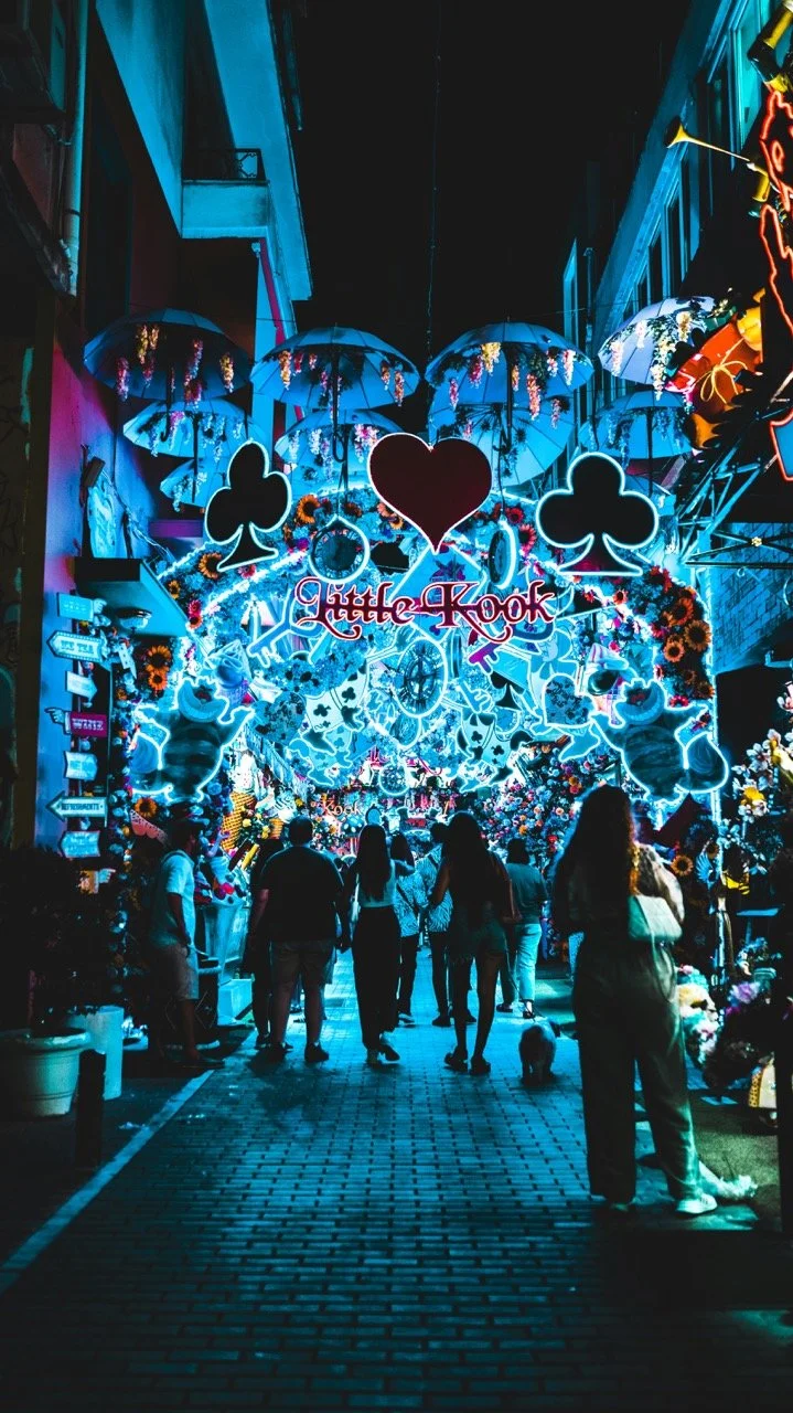 Night scene of a street in Gatlinburg decorated with neon lights, umbrellas hanging overhead, playing cards, hearts, clubs, and spades symbols, along with colorful flowers and a crowded pedestrian walkway.