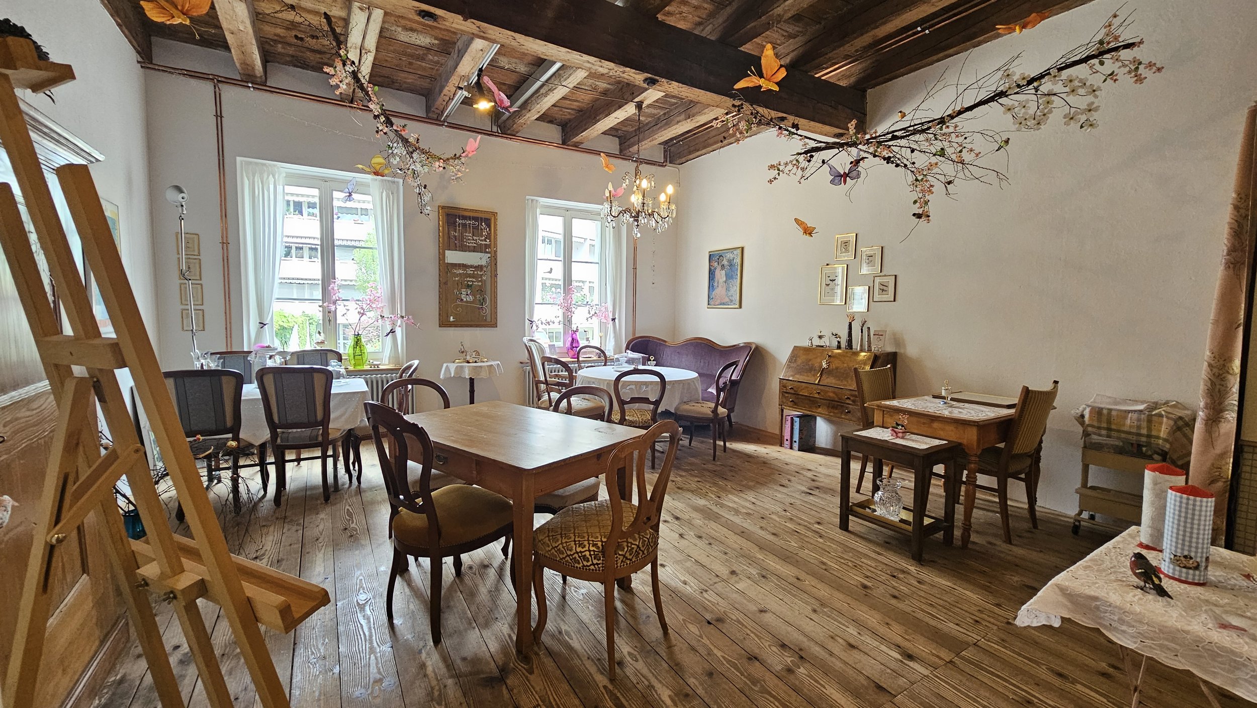 Ein rustikaler, liebevoll dekorierter Raum mit mehreren Tischen und Stühlen, viel natürlichem Licht durch Fenster, und einer Decke mit Holzbohlen. Blumen und Schmetterlingsdekoration hängen von der Decke.
