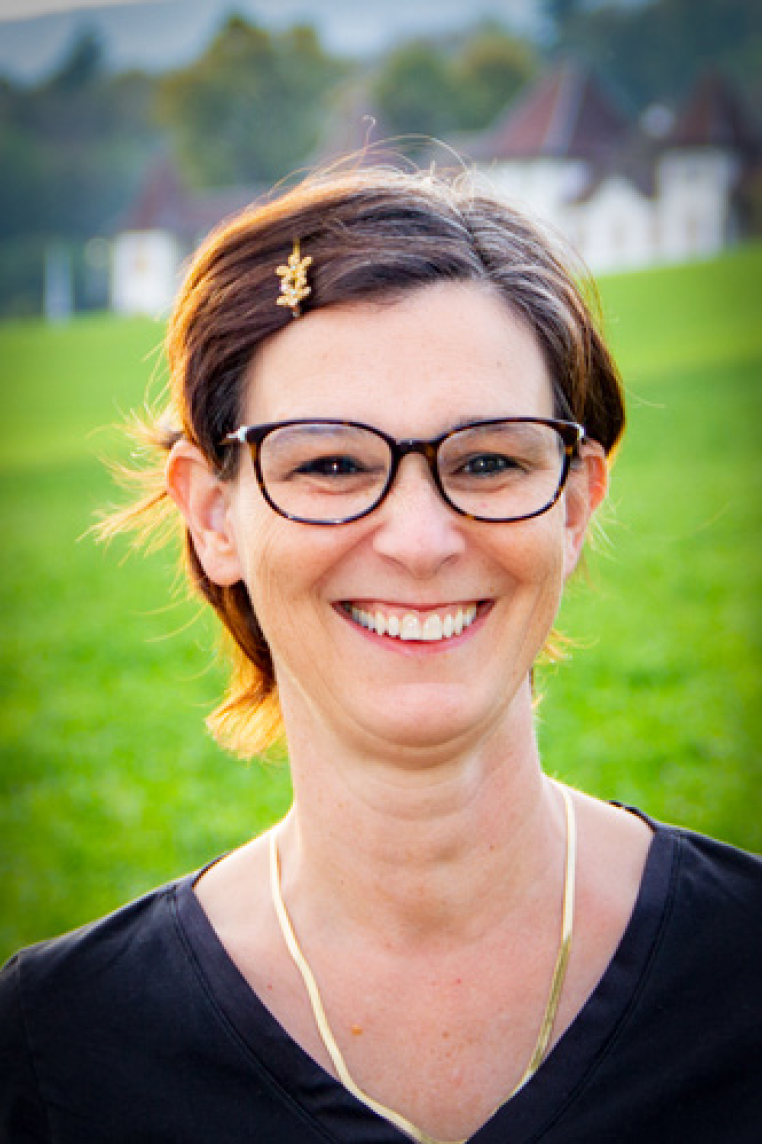 Lächelnde Frau mit Kurzhaarfrisur, Brille und schwarzem T-Shirt im Freien, grüner Hintergrund mit Bäumen und Gebäuden.