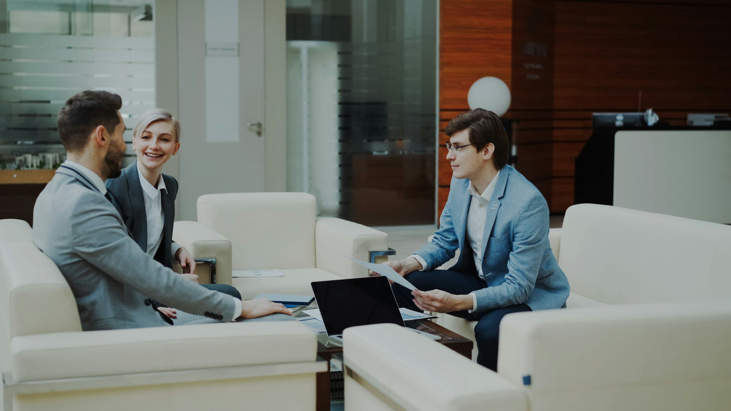 Three young professionals having a business meeting in a modern office lounge, sitting on white sofas with laptops and paperwork.