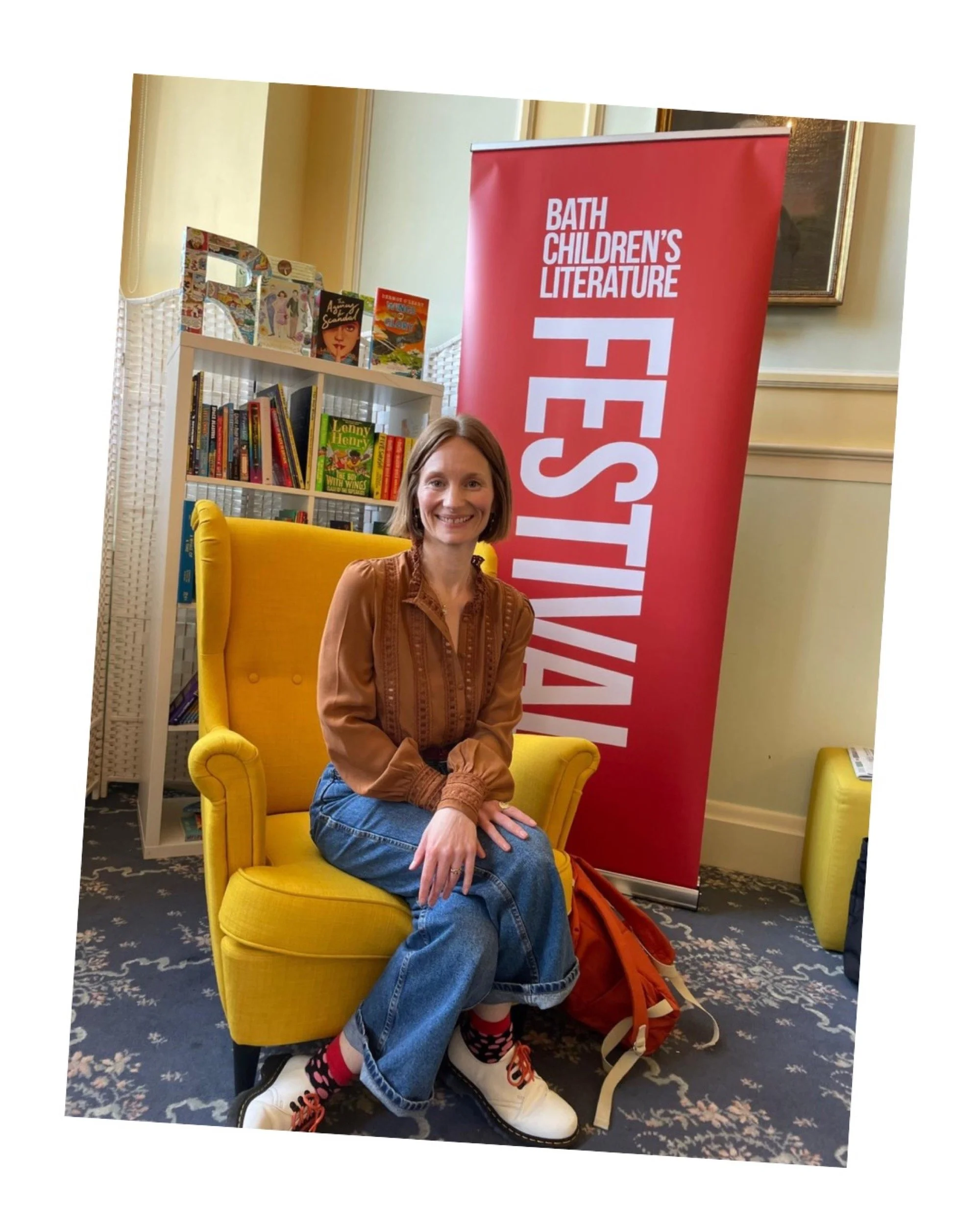 A woman sitting on a yellow armchair in front of a red banner that reads 'Bath Children’s Literature Festival.' She is smiling and wearing a brown blouse, blue jeans, and colorful socks with white shoes. There are bookshelves with children's books in
