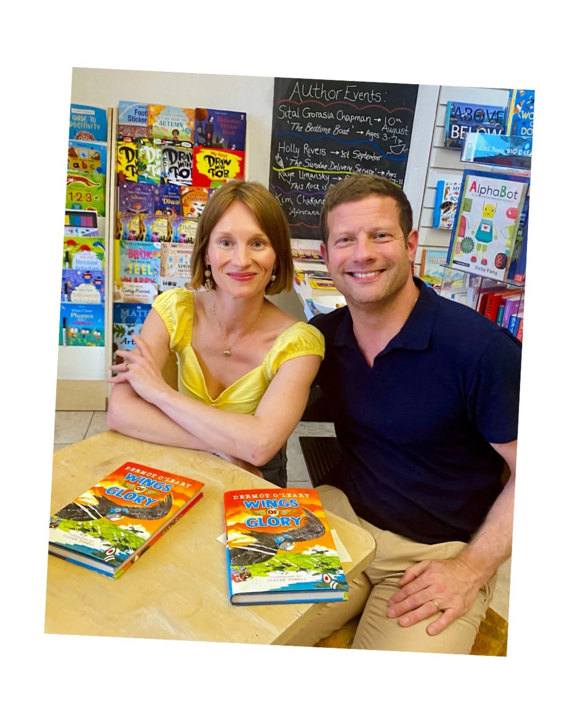 A woman with shoulder-length red hair in a yellow top and a man with short brown hair in a black shirt sit at a table with two copies of a children's book titled "Wings of Glory" by Dermot O'Leary. They are smiling at the camera inside a bookstore or