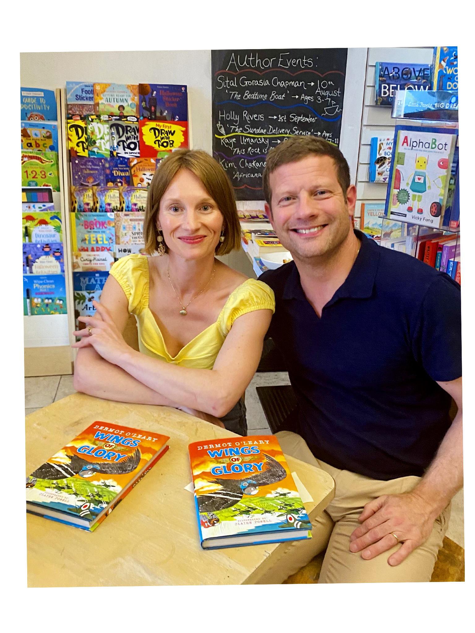 A woman and a man sitting at a table inside a bookstore or library, with copies of the book "Wings of Glory" on the table in front of them, and bookshelves filled with children's books behind them.