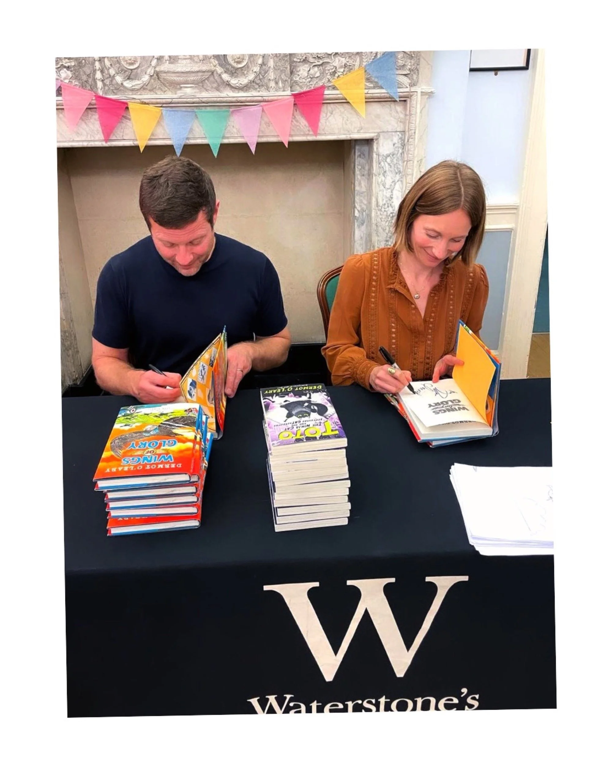 Two authors sitting at a table signing books, with stacks of their books in front of them, and a colorful bunting banner hanging above.