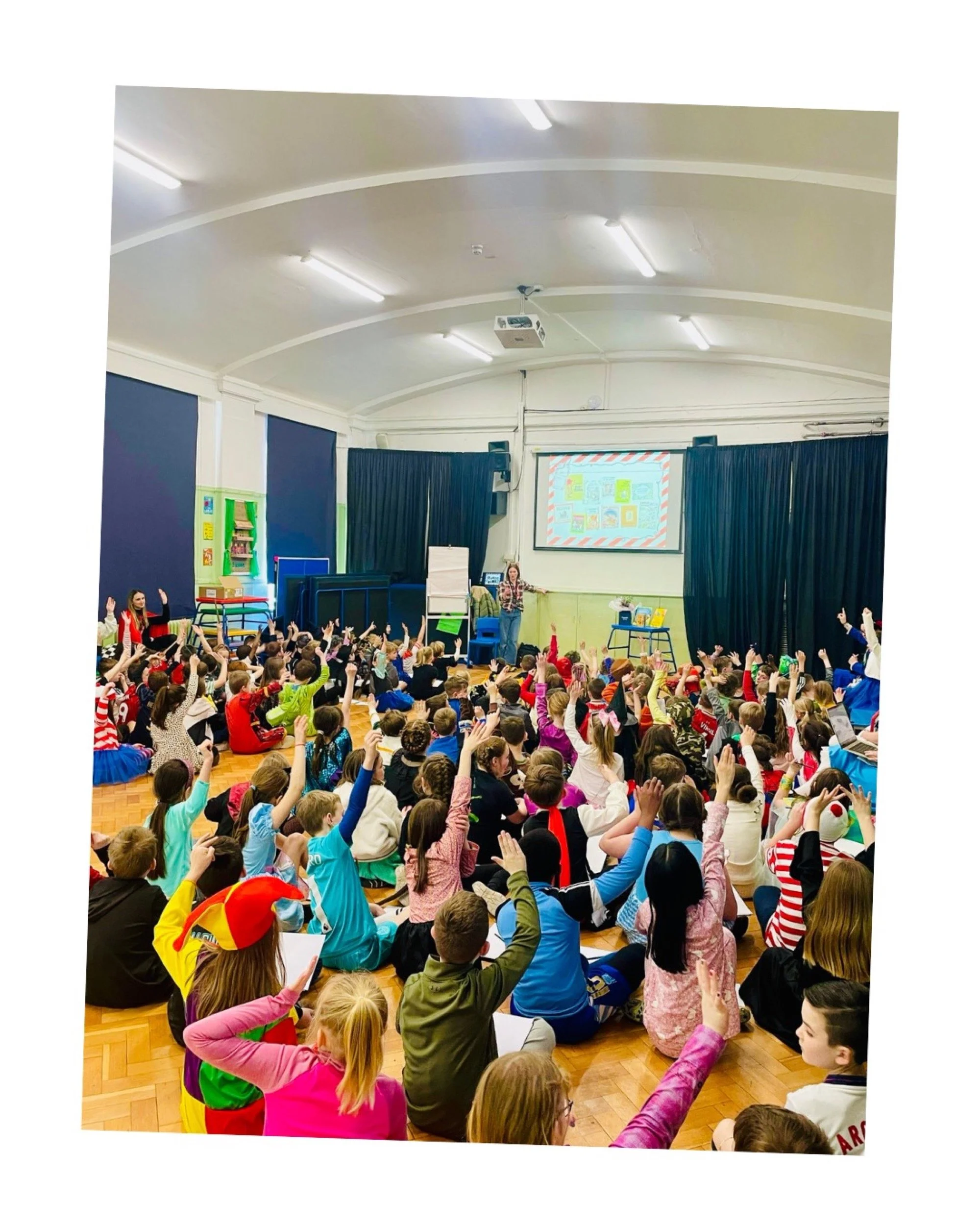 A classroom filled with young students sitting on the floor, many raising their hands, listening to a teacher at the front of the room. The classroom has a projector screen, blue curtains, and colorful decorations.
