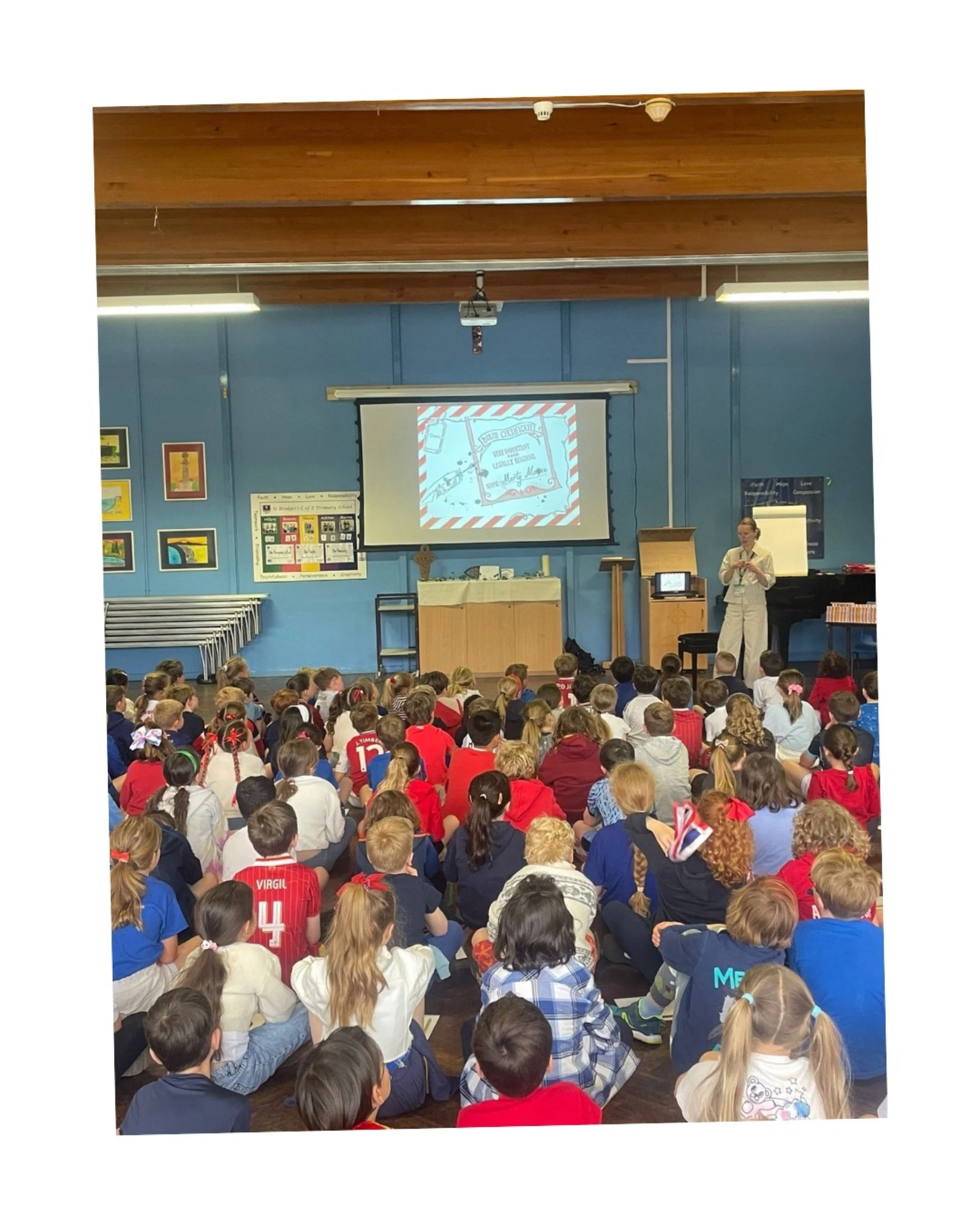 A large group of elementary school students sitting on the floor in a classroom, listening to a woman giving a presentation at the front.