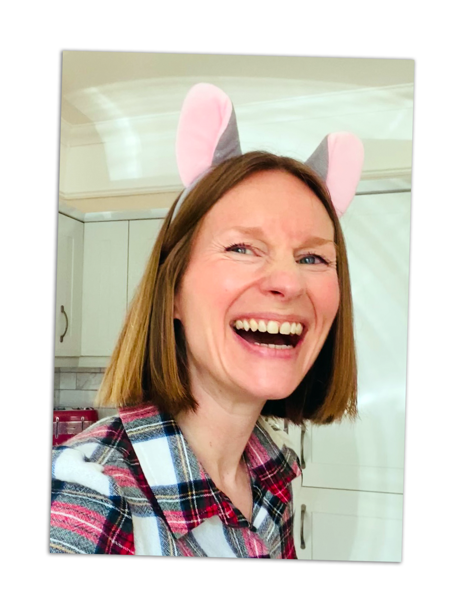 A woman with shoulder-length red hair smiling and wearing pink and gray bunny ears. She is indoors, in a kitchen with white cabinets, dressed in a red, white, and blue plaid shirt.