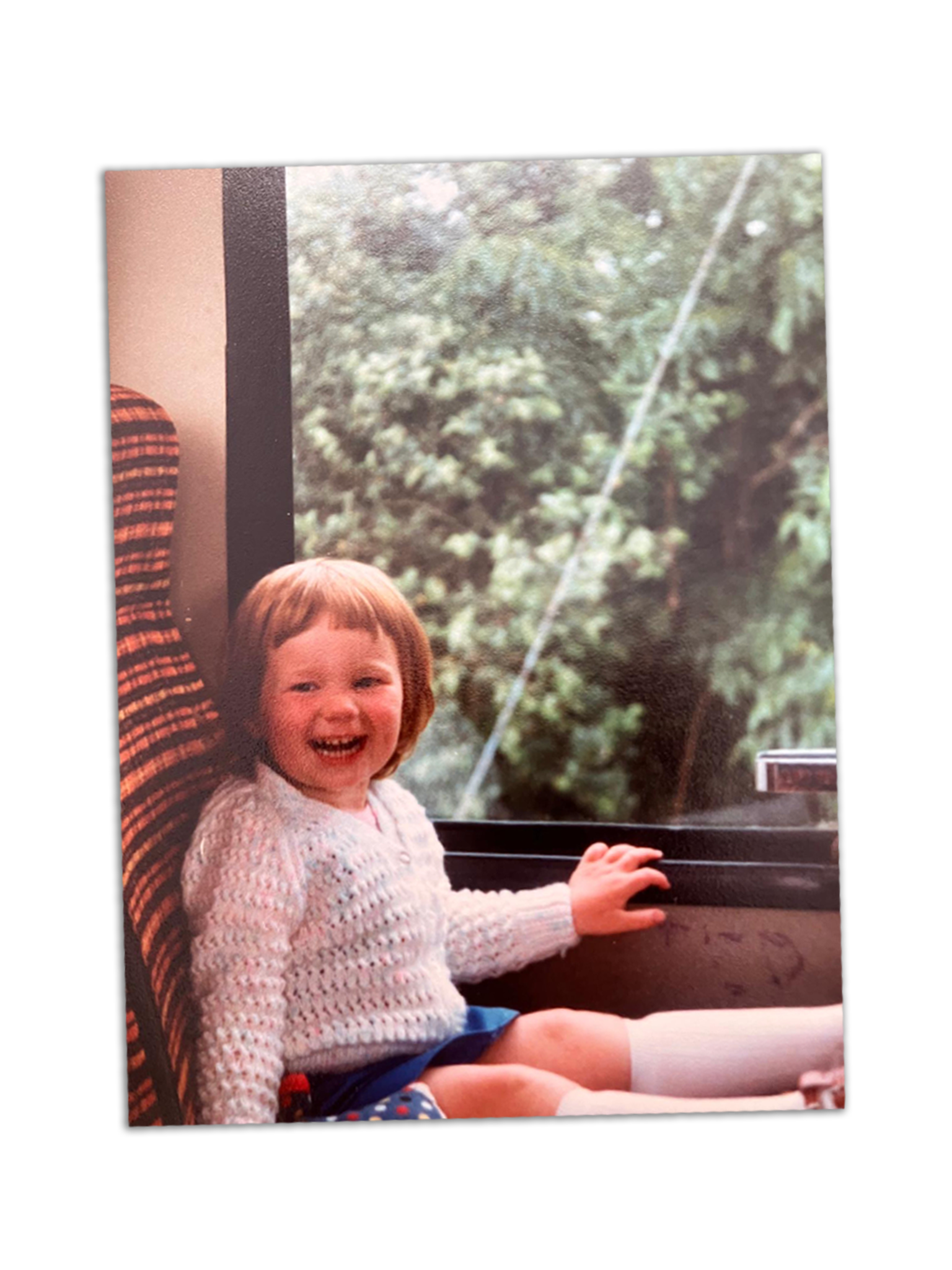 Young girl with short brown hair, smiling and laughing, sitting on a bus or train with a window showing green trees outside.