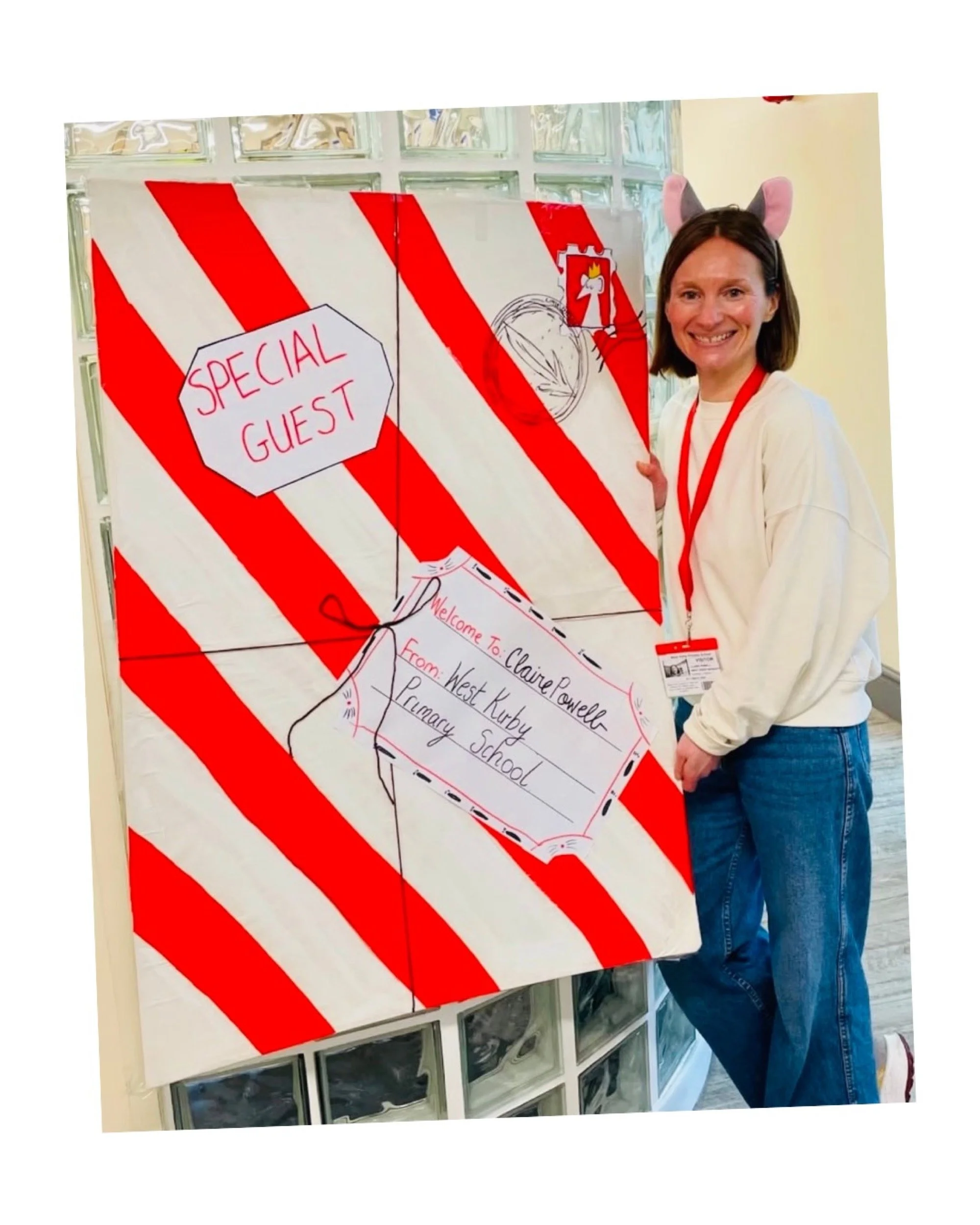 A woman with bunny ears headband smiling next to a colorful, handmade welcome sign for a special guest at a school event. The sign is decorated with red and white stripes and says "Welcome To: Claire Powell, From: West Kirby Primary School."