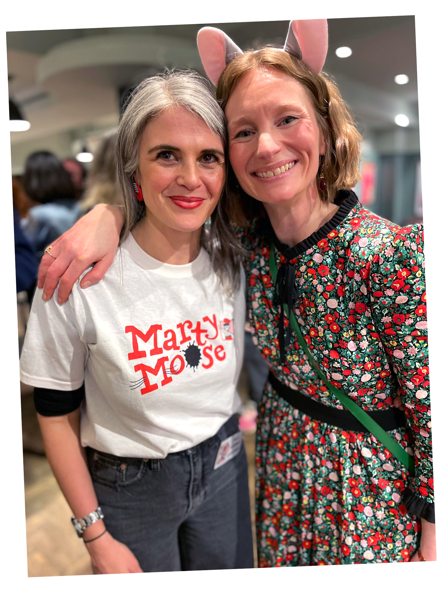 Two women, one with gray hair wearing a 'Marty Mouse' T-shirt, and the other with red hair wearing a floral dress and pink cat ears headband, smiling and standing close together in a crowded indoor setting.