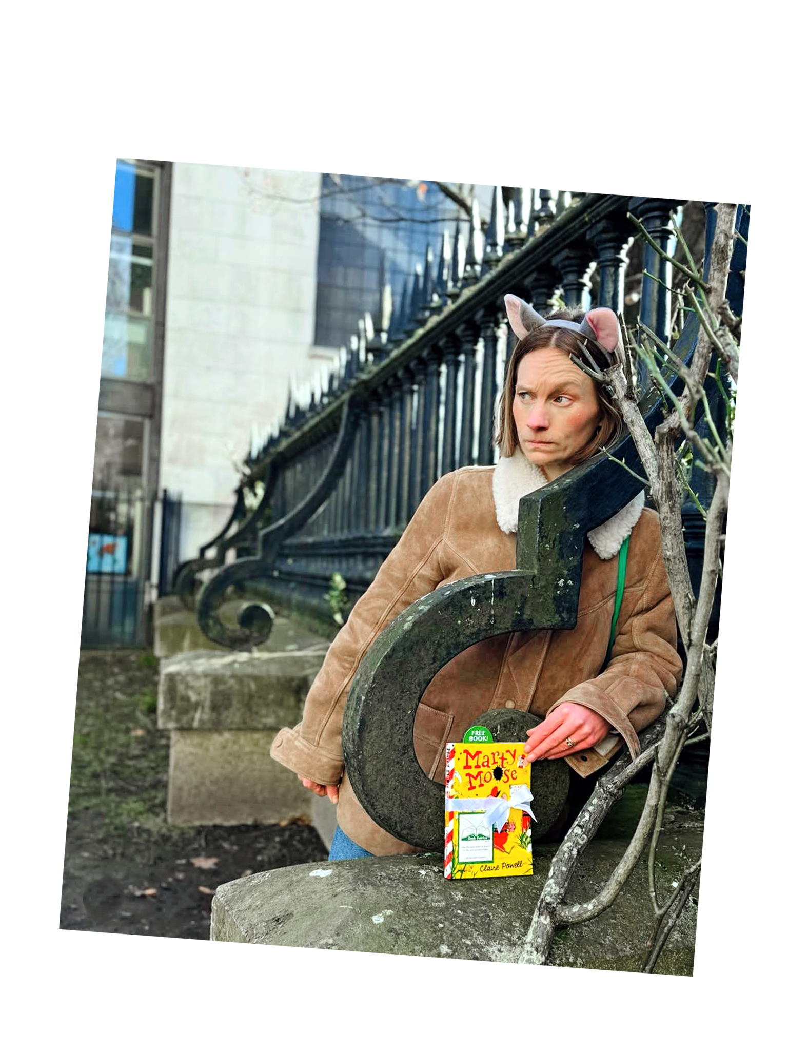 Woman dressed as a fox, wearing a headband with fox ears, sitting on a park bench with a book titled "Marty Mouse" on her lap, next to a leafless tree against a backdrop of a black iron fence and building.