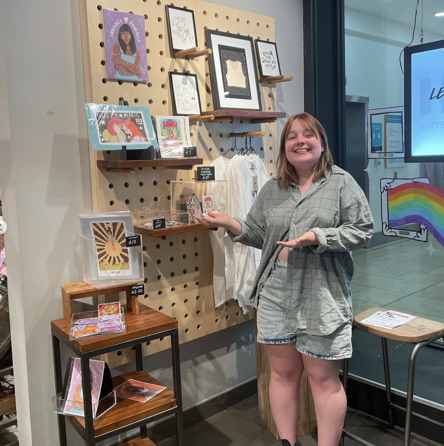 A young woman with red hair, wearing a light gray plaid button-up shirt and matching shorts, standing inside a store with a pegboard wall filled with framed artwork and small items. She is smiling and holding a small item in her right hand.