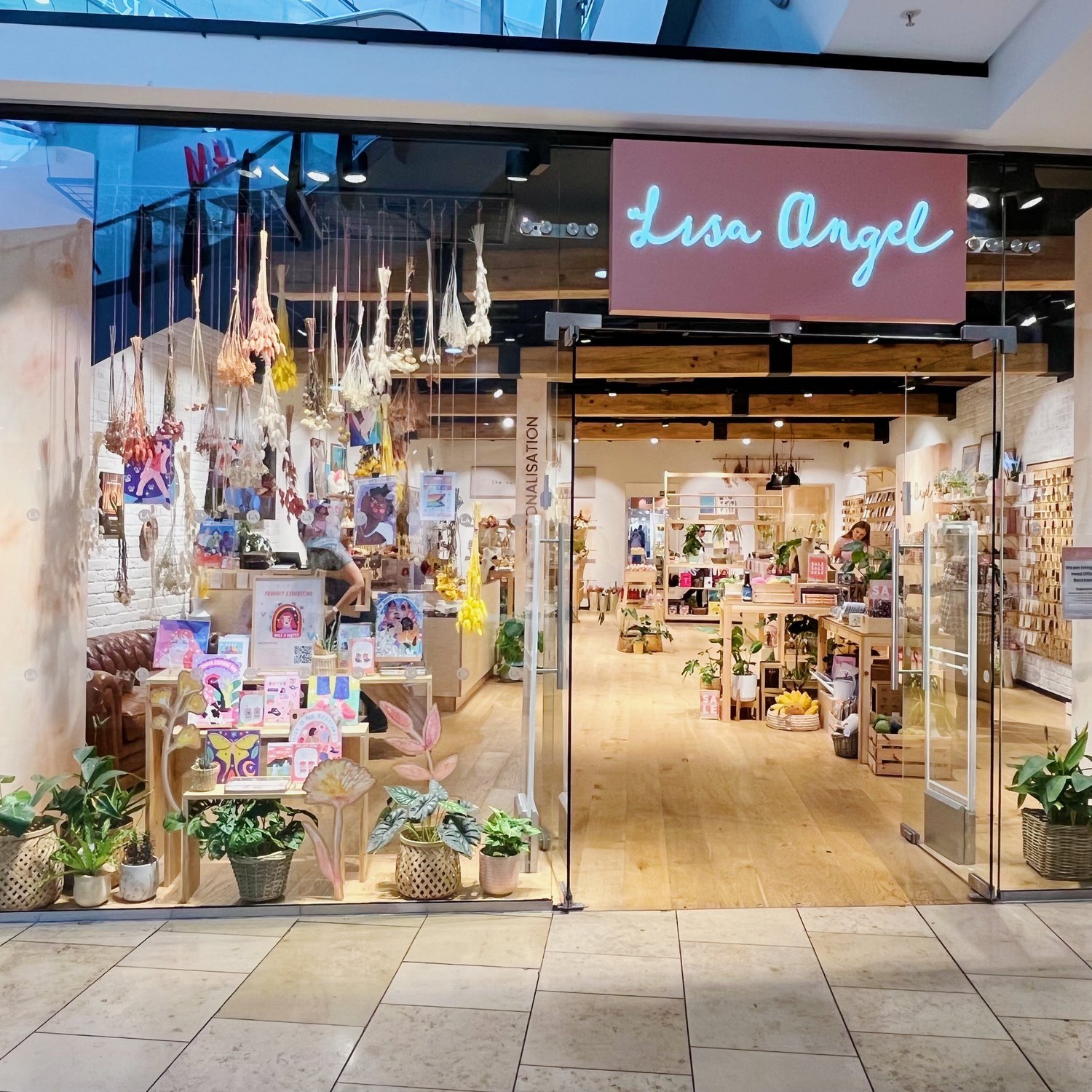 Front view of a retail store named "Lisa Angel" with a glass front door, interior decorated with plants, shelves, and hanging crafts, inside a shopping mall.