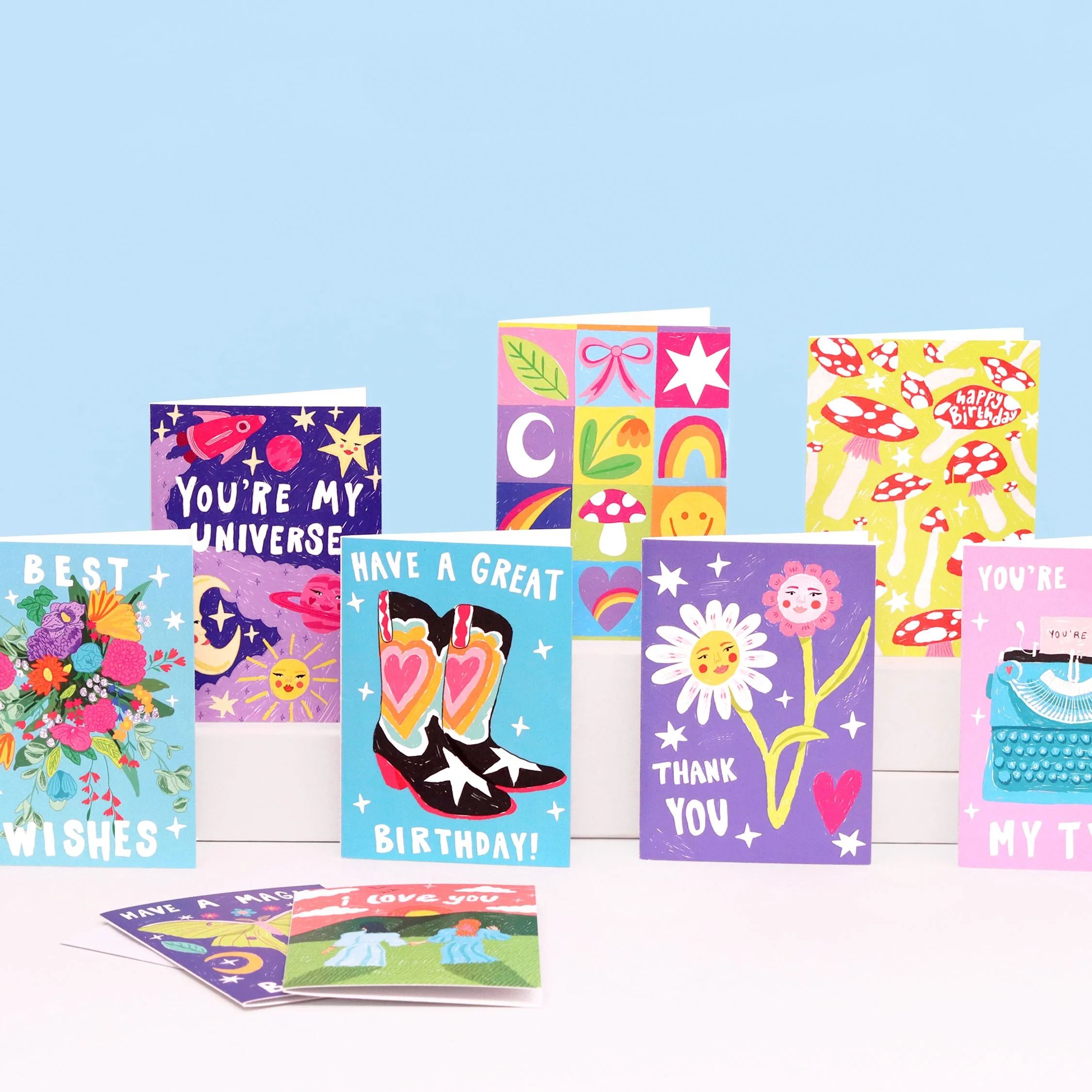 Colorful birthday and greeting cards with various illustrations and messages, arranged on a white surface against a light blue background.